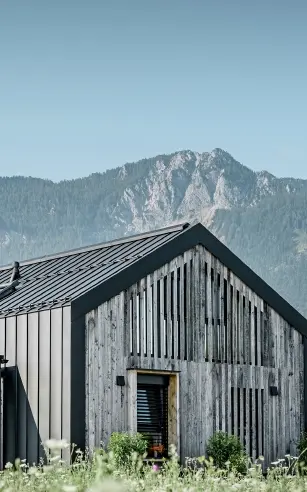 Moderner, funktionaler Wohnbau mit einer Kombination aus Holzfassade und PREFA Band- und Blechmaterial für das Dach. Die klare Architektur wird durch die elegante, metallische Dach- und Fassadenverkleidung unterstrichen, die ideal für anspruchsvolle Projekte ist. Das Bild veranschaulicht die vielseitigen Anwendungsmöglichkeiten der PREFA Produkte im Bereich Bänder und Bleche.