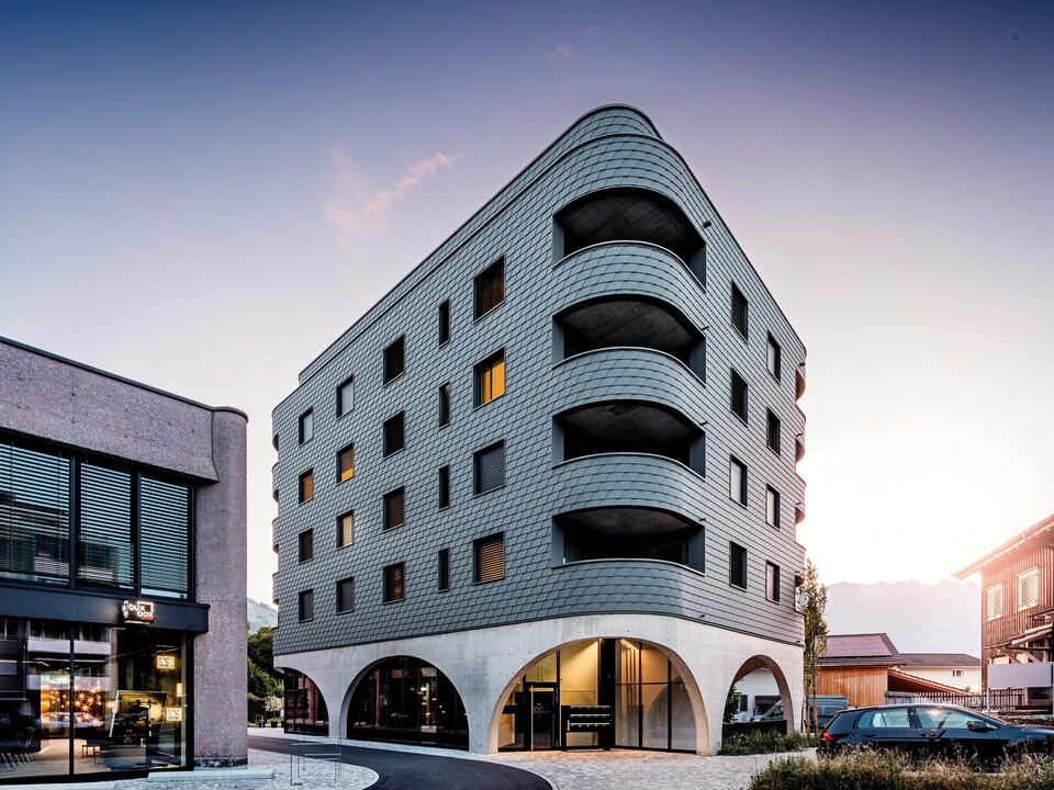 Modernes Mixed-Use-Gebäude in Buchs mit markant gerundeter, dreieckiger Form und durchgehender Fassade aus PREFA Wandschindeln in P.10 Hellgrau. Die schuppenartige Aluminiumhaut umhüllt das Gebäude wie eine Reptilienstruktur und betont die fließenden Rundungen sowie die tief eingeschnittenen Balkone. Über dem massiven Sockel mit eleganten Betonarkaden wirkt die leichte, witterungsbeständige PREFA Fassade besonders ausdrucksstark. Präzise verlegte Aluminiumschindeln sorgen für eine homogene, langlebige und wartungsarme Gebäudehülle mit starkem Wiedererkennungswert.