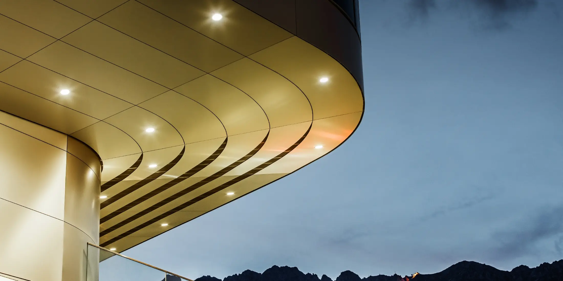 The image shows an impressive architectural detail view of a PREFA PEMA composite panel made of brushed aluminium. The panel is gently curved and flows in elegant, flowing lines, which are particularly accentuated by targeted lighting in the evening. The clear structure and the elegant aluminium look of the panel complement the modern design of the façade and offer an elegant, contemporary solution for high-quality architecture.