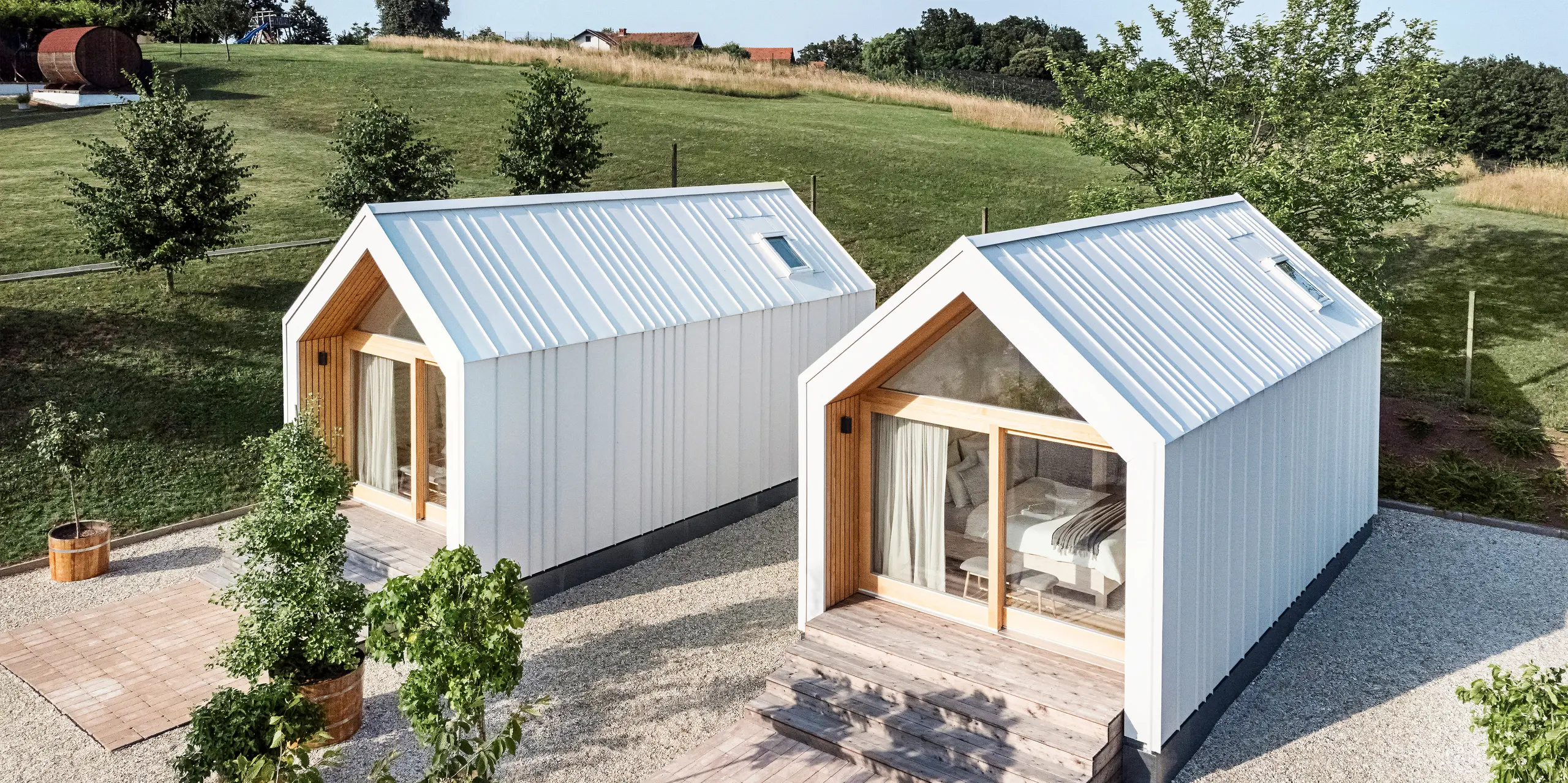 View of the front of the two tiny houses in the 'Pri Momi' holiday complex in Kančevci, Slovenia. The micro houses were built using a timber frame construction and finished with a robust PREFALZ standing seam roof and façade cladding in P.10 Pure White. The modern, minimalist buildings combine a durable yet aesthetic aluminium shell with large glass fronts and natural wood, embedded in a green landscape. The generous wooden steps lead up to the entrances, creating a cosy and inviting atmosphere.
