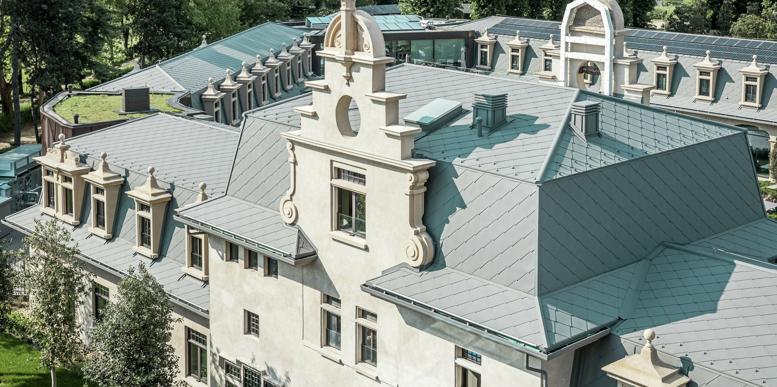 Detaillierte Dachansicht des Thermalparks „De Montel – Terme Milano“ mit präzise ausgeführter Eindeckung aus PREFA Dachrauten 44 × 44 in P.10 Hellgrau. Die komplexe Dachgeometrie mit zahlreichen Gauben, Gesimsen, Giebeln und Lüftungselementen wird handwerklich meisterhaft gelöst. Harmonisch ergänzt wird die historische Architektur durch moderne Elemente wie ein begrüntes Flachdach und die nahtlos integrierte Photovoltaikanlage im Hintergrund.
