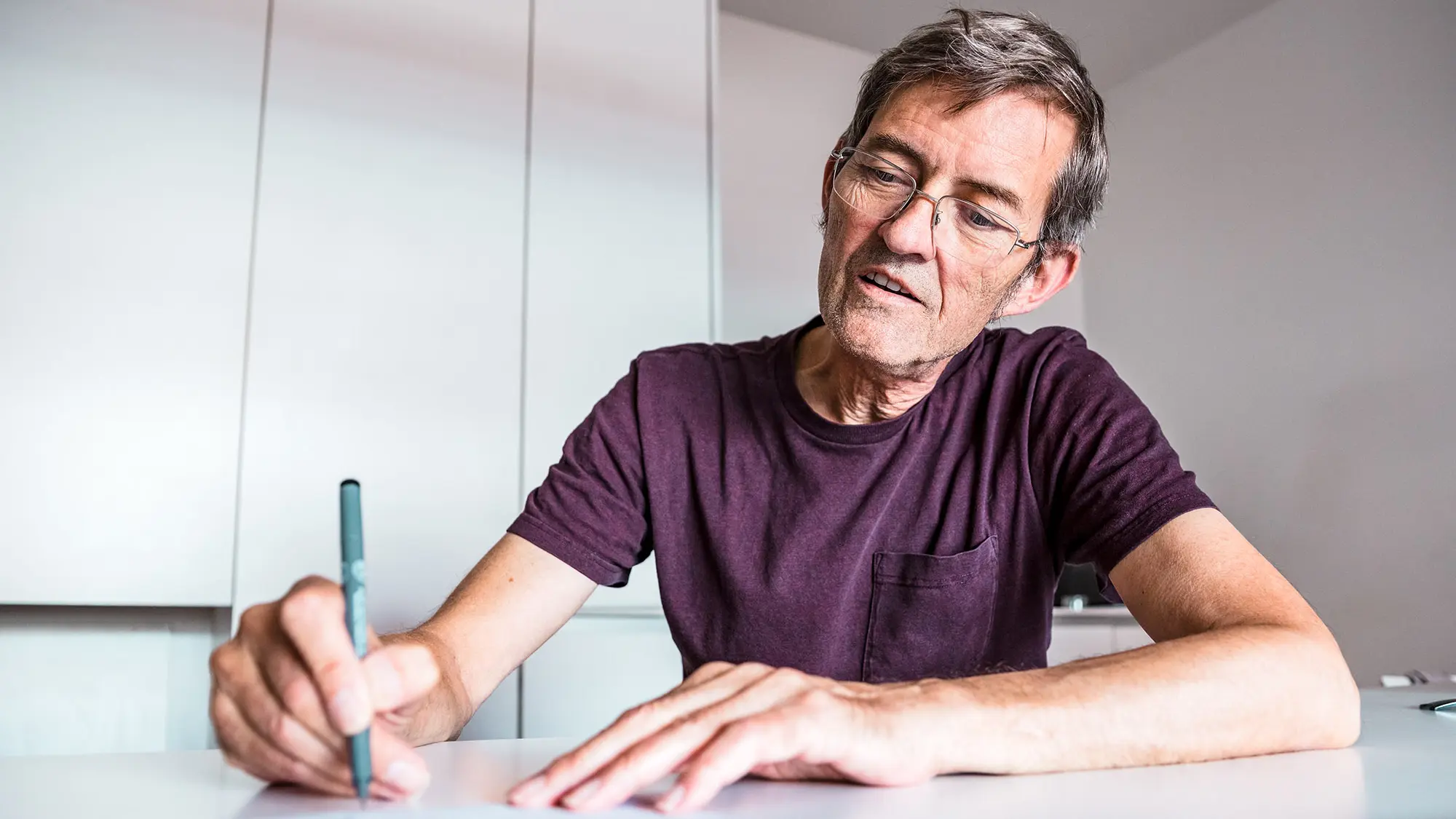 Architect Dominique Cornaert sitting at a white table and drawing; behind him, white walls as well as a white cupboard can be seen.