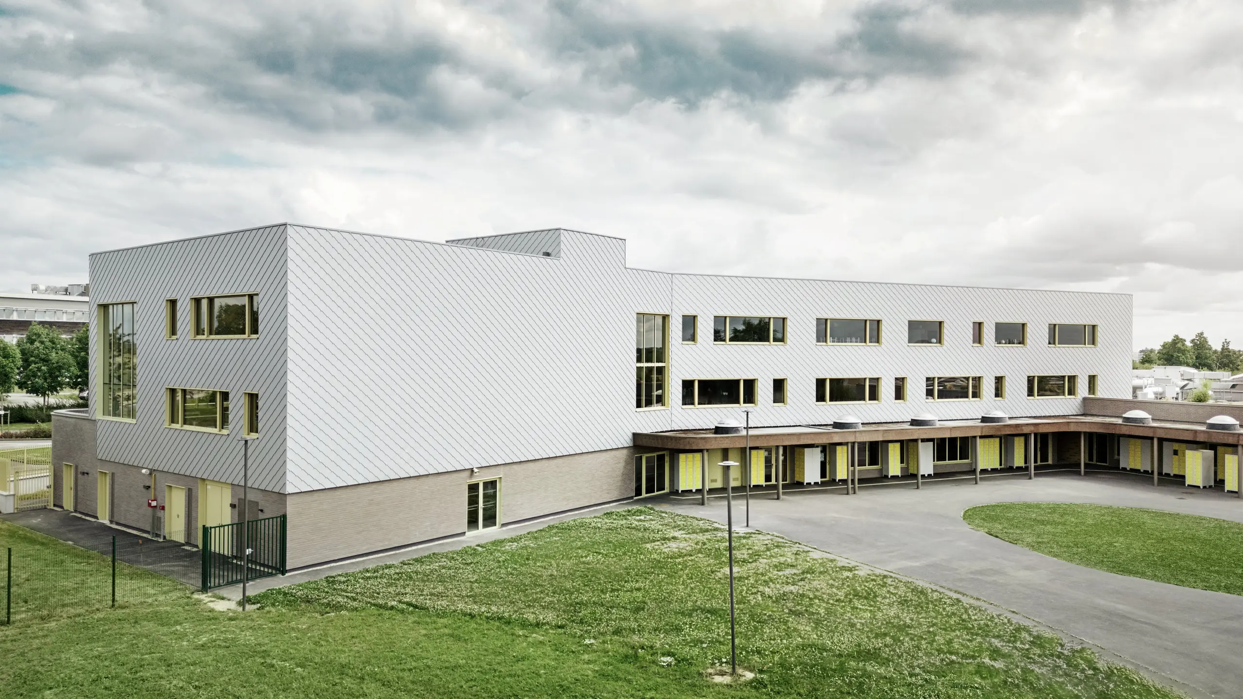 Das Bild zeigt die Rückseite des Collège Léontine Dolivet. Im Vordergrund befindet sich ein großzügiger, gepflasterter Schulhof mit klaren Linien und offenen Flächen, der sich ideal für Pausen und Freizeitaktivitäten eignet. Mehrere Grünflächen lockern die eintönige Umgebung auf. Im Hintergrund erhebt sich ein modernes Schulgebäude mit einer markanten Aluminiumfassade aus silbermetallischen PREFA-Wandelementen, die das Licht reflektieren und so eine dynamische Wirkung erzeugen. Zahlreiche große Fenster lassen viel Licht ins Innere. Die Aufnahme entstand tagsüber bei leicht bewölktem Himmel, wodurch die Metalloberfläche besonders gut zur Geltung kommt.