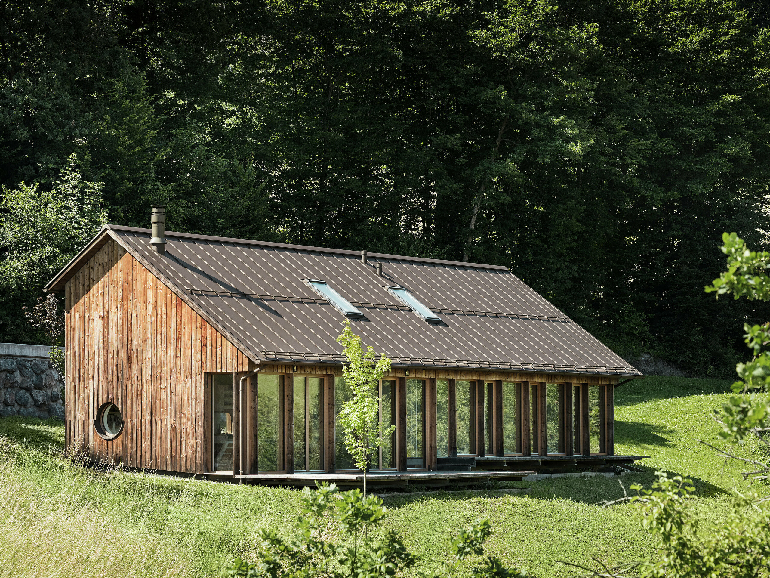 Ferienhaus in Holzbauweise mit großflächiger Glasfassade und hochwertigem PREFALZ Dach in P.10 Braun – harmonisch eingebettet in die grüne Hügellandschaft von Vinje, Slowenien. Das widerstandsfähige Aluminiumdach überzeugt durch Langlebigkeit, Korrosionsschutz und moderne Ästhetik. Integrierte Dachfenster sorgen für natürliches Licht und energieeffizientes Wohnen. Das Holzhaus wird von zahlreichen Bäumen eingerahmt.