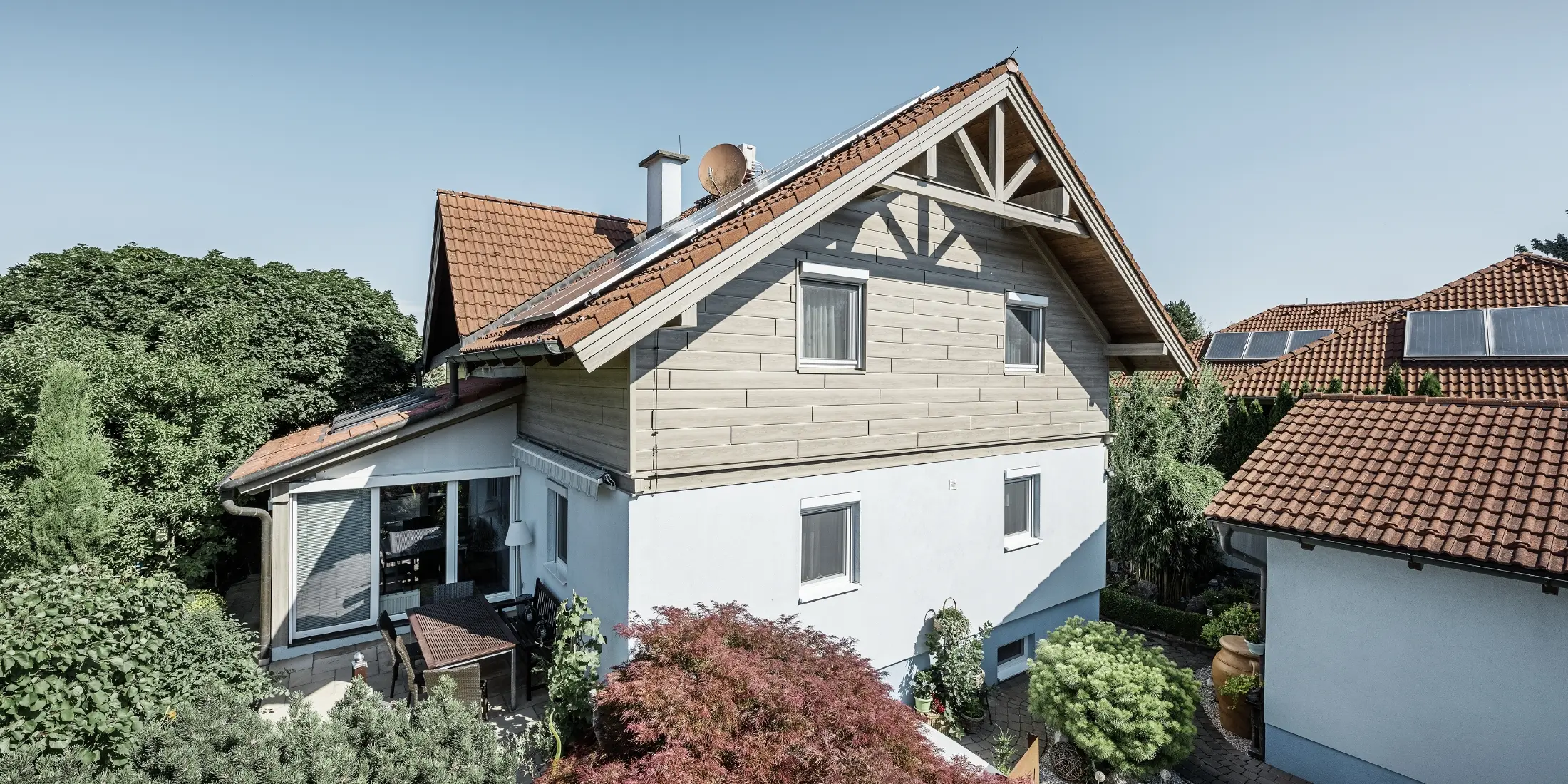 Detached house with a gable roof and red roofing, combined with a modern façade design. The upper gable is clad with beige-grey aluminium façade panels from PREFA, while the ground floor has a white plaster façade. Solar panels are installed on the roof. The house is set in a green garden with a terrace, garden furniture and lush planting, surrounded by other residential buildings with similar roof architecture.