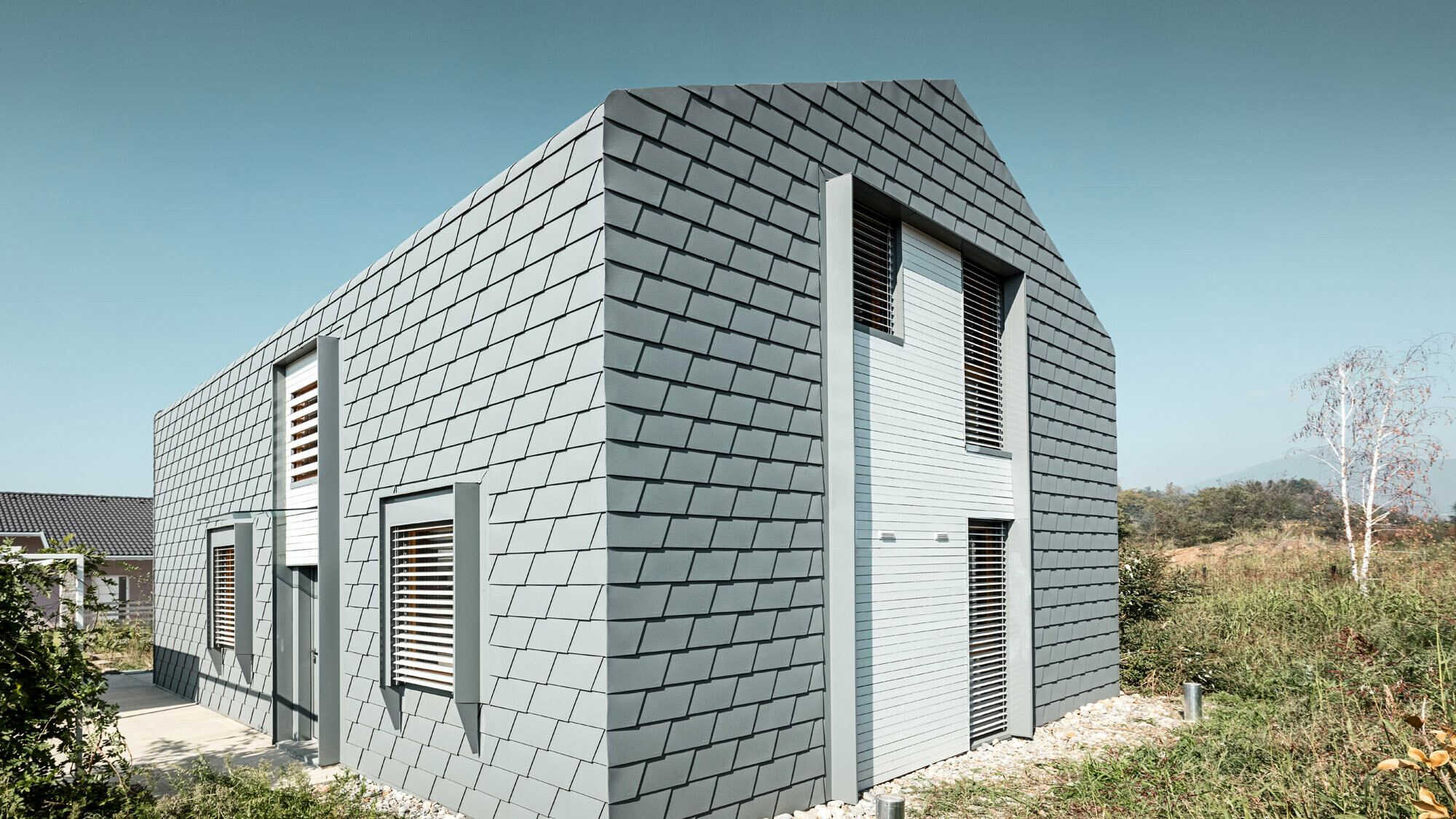 Three-quarter view of the house with green areas, the continuous shingle skin and the windows with external blinds are clearly visible.