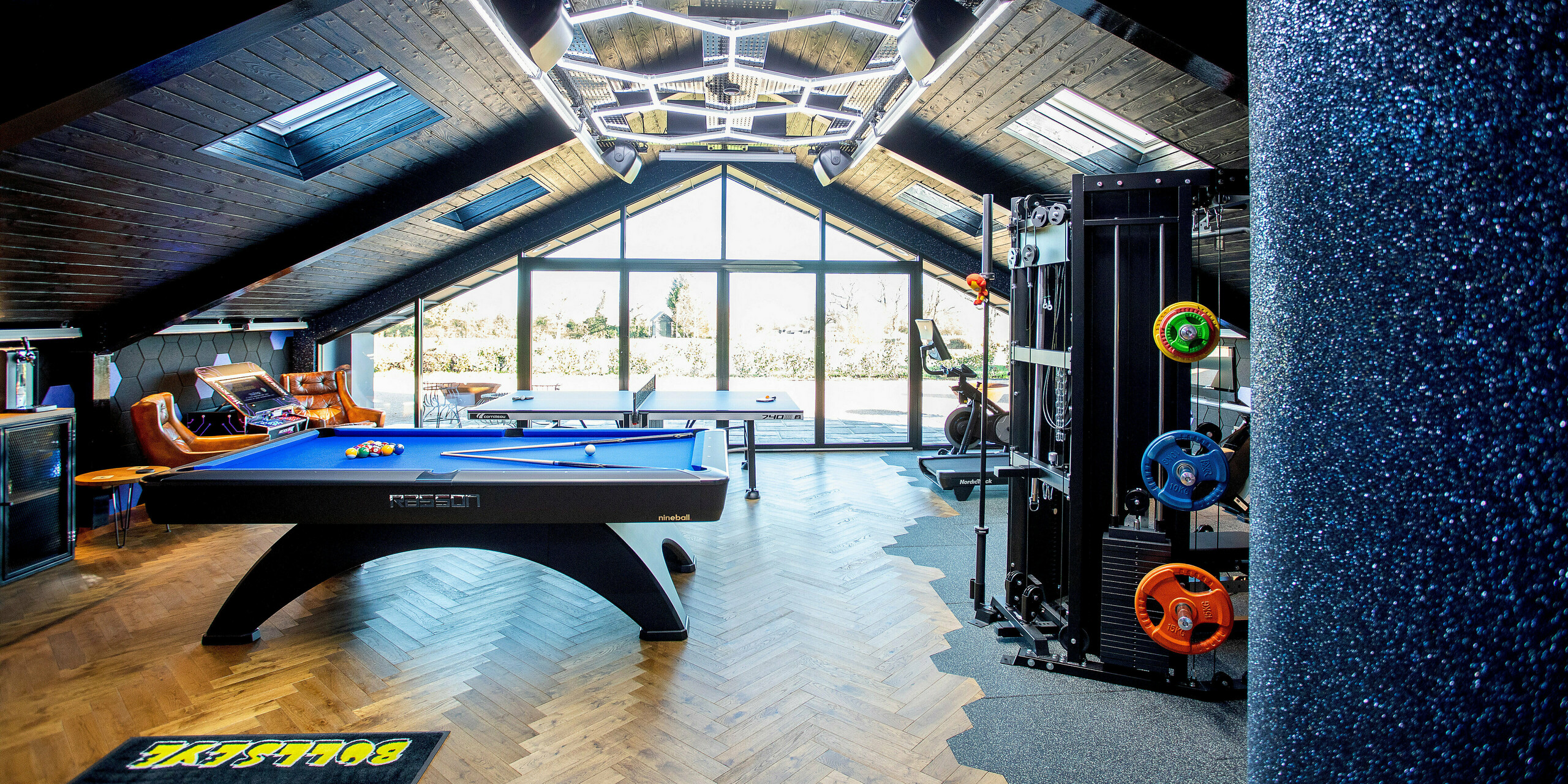 Interior view of the modern leisure building featuring a large glass façade and striking roof structure. The multifunctional space combines a pool table, table tennis, fitness equipment and arcade elements, all framed by the clean architectural lines of the PREFA aluminum roof. Premium materials, dynamic lighting and parquet flooring create an energetic, inviting environment.