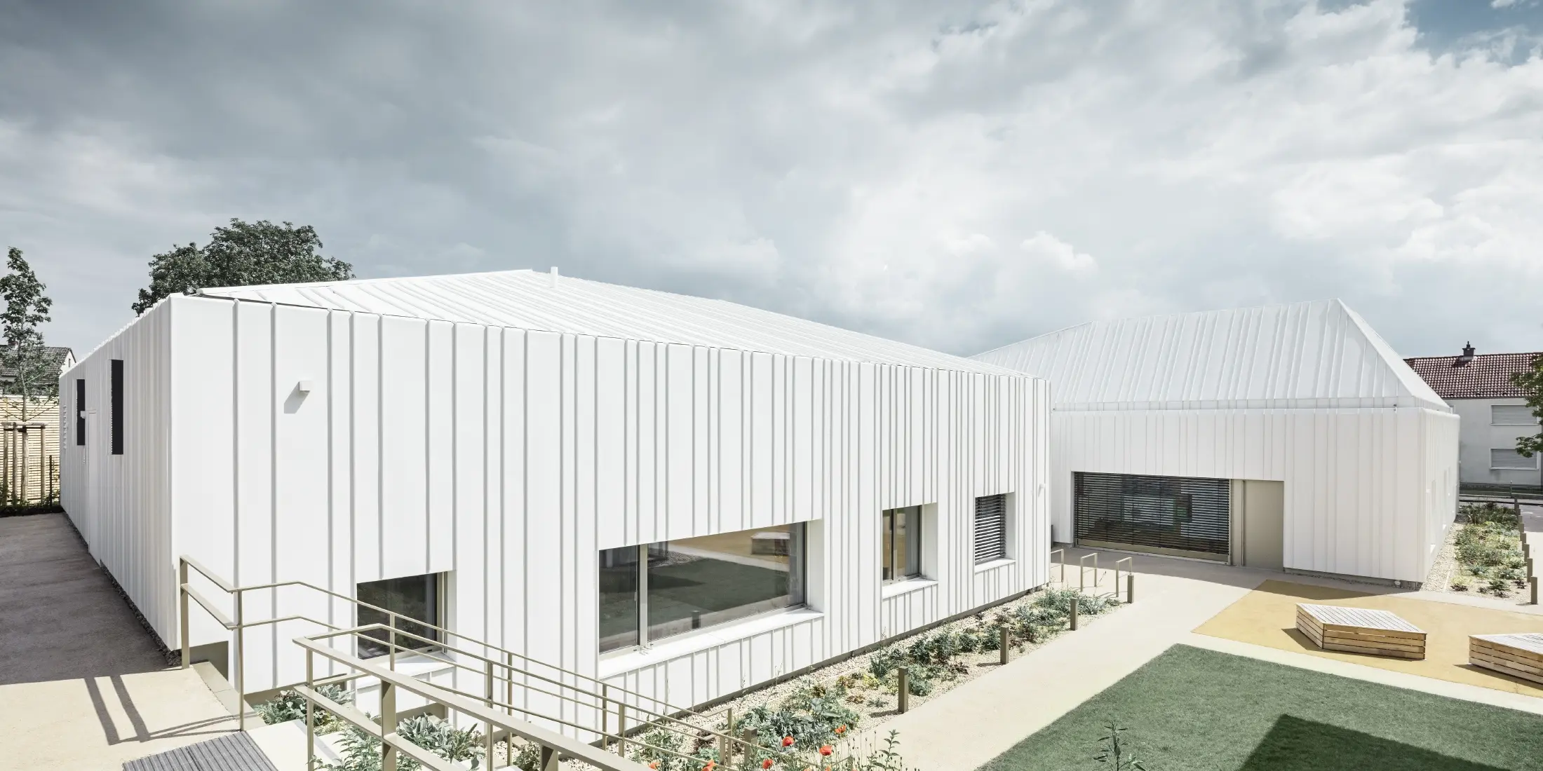 Modernes Kindergartengebäude mit klarer, geometrischer Architektur und Fassade aus weißen Aluminiumprofilen. Die Anlage besteht aus mehreren verbundenen Baukörpern mit flach geneigten Dächern. Große Fenster sorgen für viel Tageslicht, während der Außenbereich mit Wegen, Beeten und Sitzbänken gestaltet ist. Der Himmel ist teils bewölkt und verleiht der Szene eine ruhige Atmosphäre.