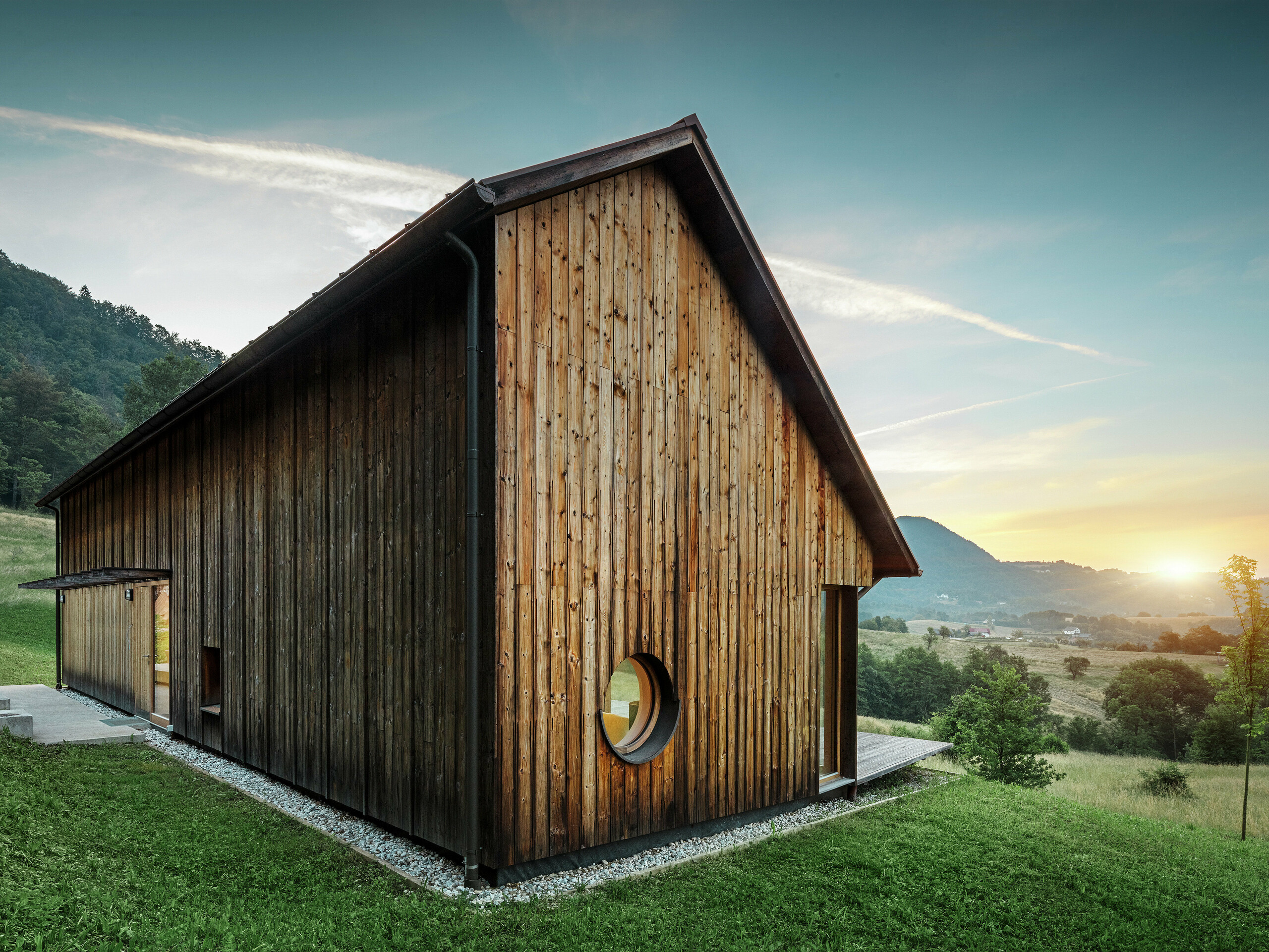 Dieses stimmungsvolle Foto zeigt ein Holzhaus in Vinje bei Sonnenuntergang mit einem robusten PREFALZ Aluminiumdach in der Farbe P.10 Braun. Das witterungsbeständige Stehfalzdach von PREFA ist langlebig, erfordert nur geringen Wartungsaufwand und fügt sich perfekt in die natürliche Umgebung ein. Die Kombination aus klarer Formensprache, Holzfassade und runder Fensteröffnung betont den architektonischen Minimalismus. Das schlichte Satteldach besticht durch seine bewusste Asymmetrie, die ihm eine ganz besondere Note verleiht.