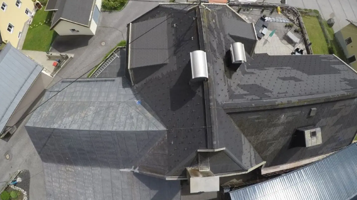 Aerial view of an old roof with many details – channels, dormers and chimneys. The old roof has been repaired multiple times and is stained.