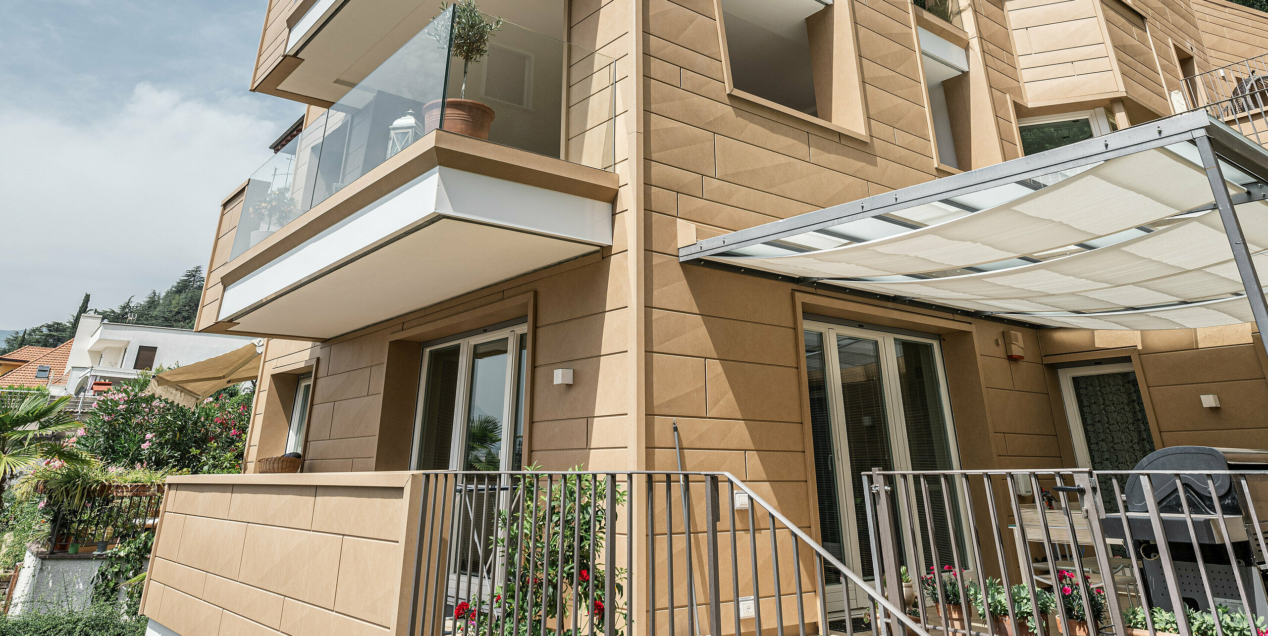 Blick aufwärts an der Fassade der Villa Laura, kunstvoll gestaltet mit PREFA Siding.X in Sandbraun, die moderne Architektur und Funktionalität in einer langlebigen Aluminiumhülle vereint. Überhängende Balkone und große Fensterflächen öffnen das Gebäude zum Himmel, während die feine Textur der Fassadenpaneele und deren natürliche Farbgebung das Gebäude optisch erden und in die mediterrane Landschaft Südtirols einbetten.