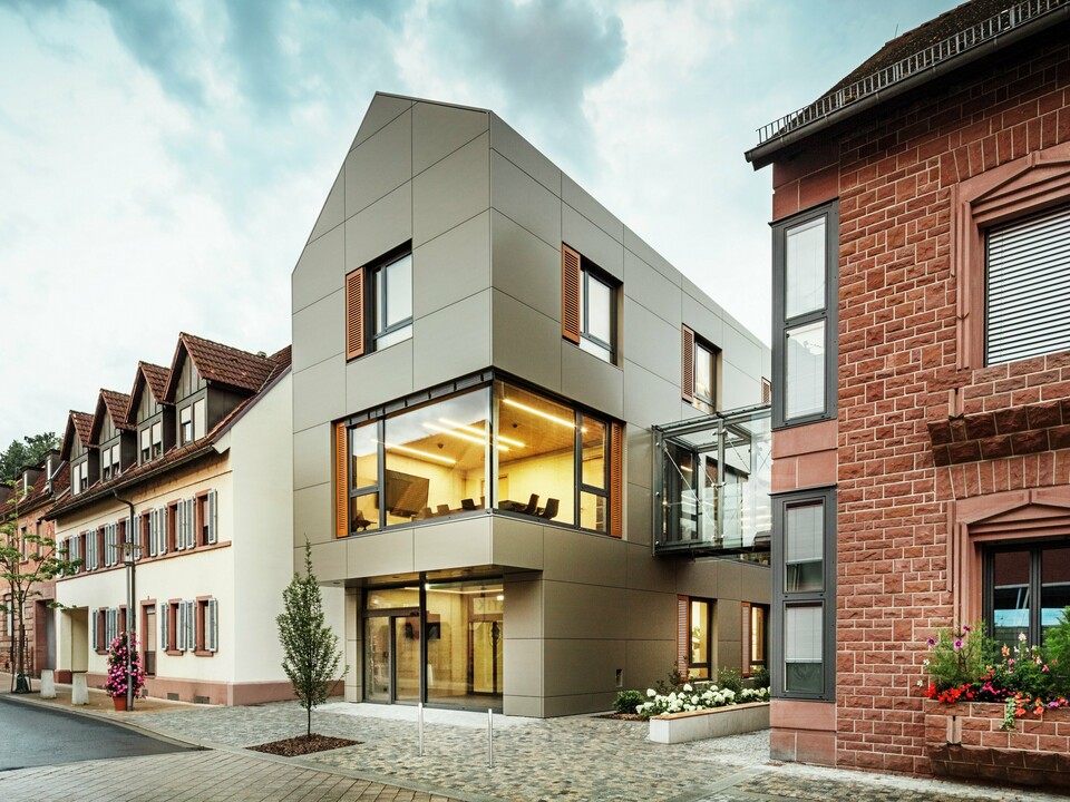 The image shows the modern town hall in Alzenau, which is clad in aluminium composite panels from PREFA in a warm bronze tone. The façade is interrupted by large windows that offer a view of the interior, which is illuminated by warm light. The glass walkway on the right-hand side of the picture connects the building to a neighbouring traditional brick building, creating an exciting contrast between modern and historic architecture.