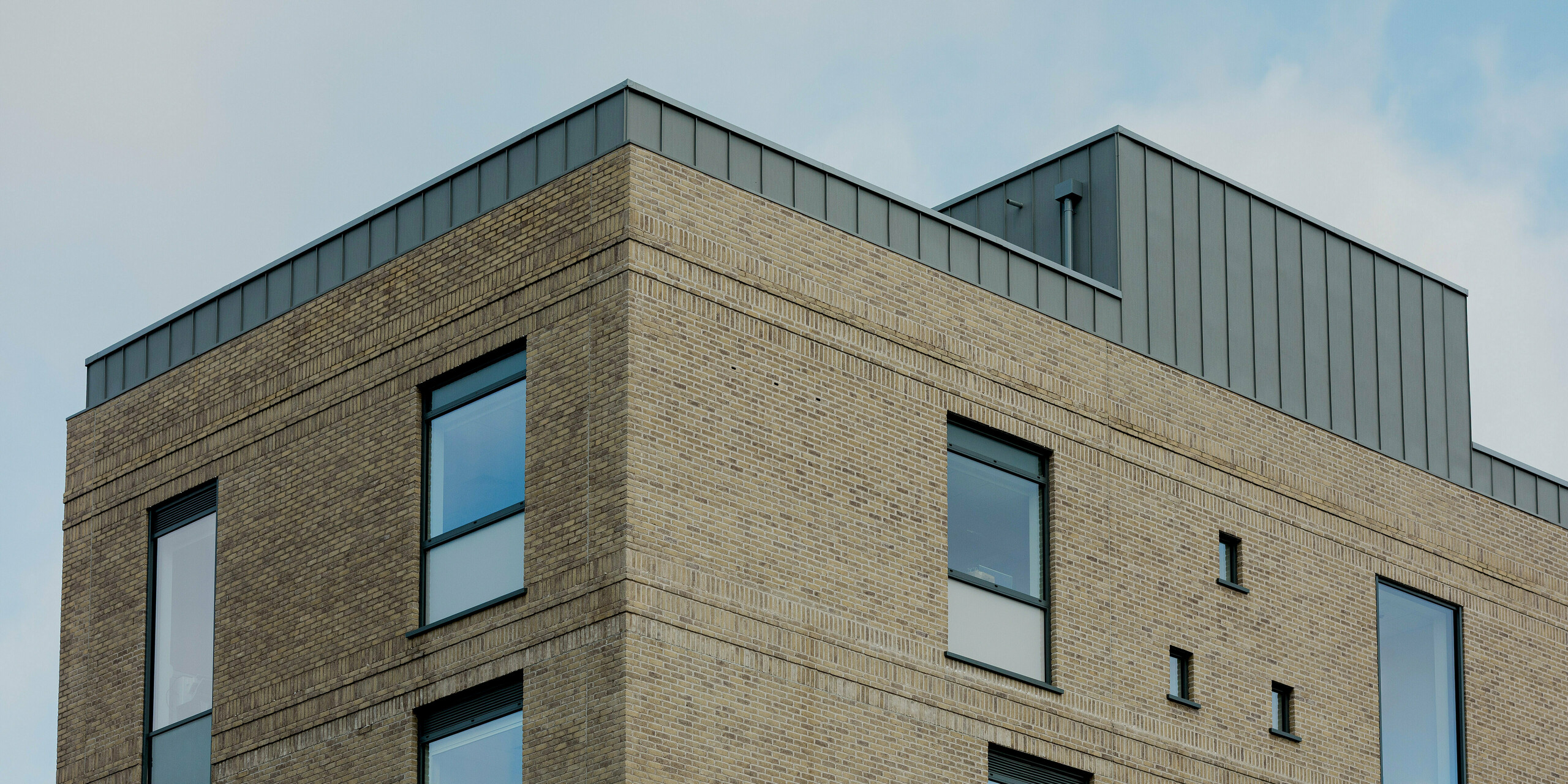 A sharp building corner of the Northern Regional College in Ballymena is defined by light brickwork and precisely proportioned windows. Along the roof edge and set-back upper volume, PREFALZ standing seam aluminium panels in patina grey form a distinctive vertical crown. The refined metal cladding contrasts subtly with the textured masonry and reinforces the building’s geometric clarity. PREFA aluminium delivers durability, weather resistance and a timeless architectural finish for contemporary educational buildings.