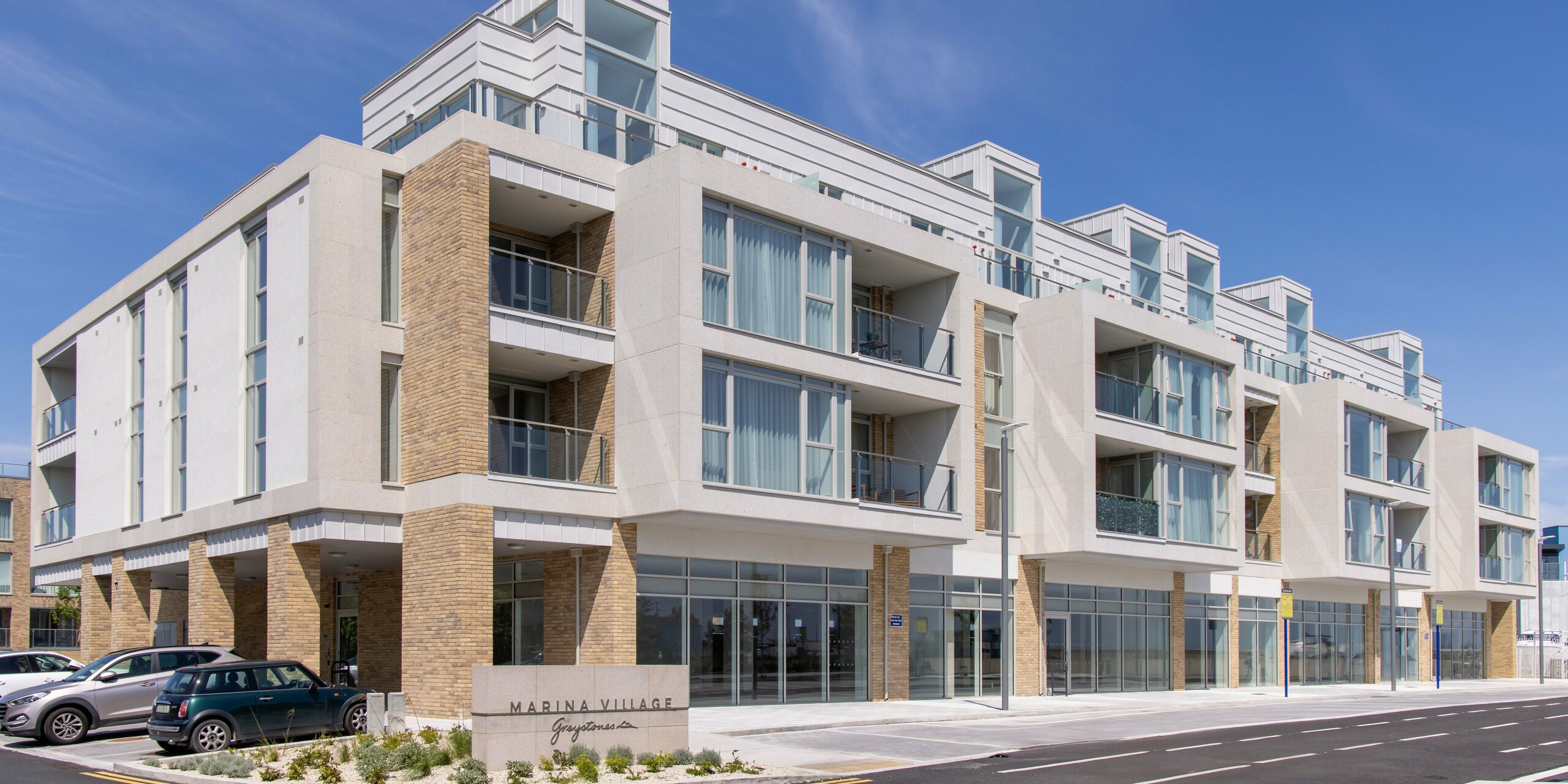 Marina Village Greystones in Wicklow, Ireland - Modern residential development on the waterfront with direct access to the marina. The development is clad in approximately 2,000 m² of robust PREFALZ in P.10 Prefa White and P.10 Light Grey. The stylish architecture features large glass fronts and balconies, ideal for exclusive living by the sea. The picture shows the entrance to one of the apartment blocks. The blue sky in the background enhances the light colours of the high quality sheet metal cladding.