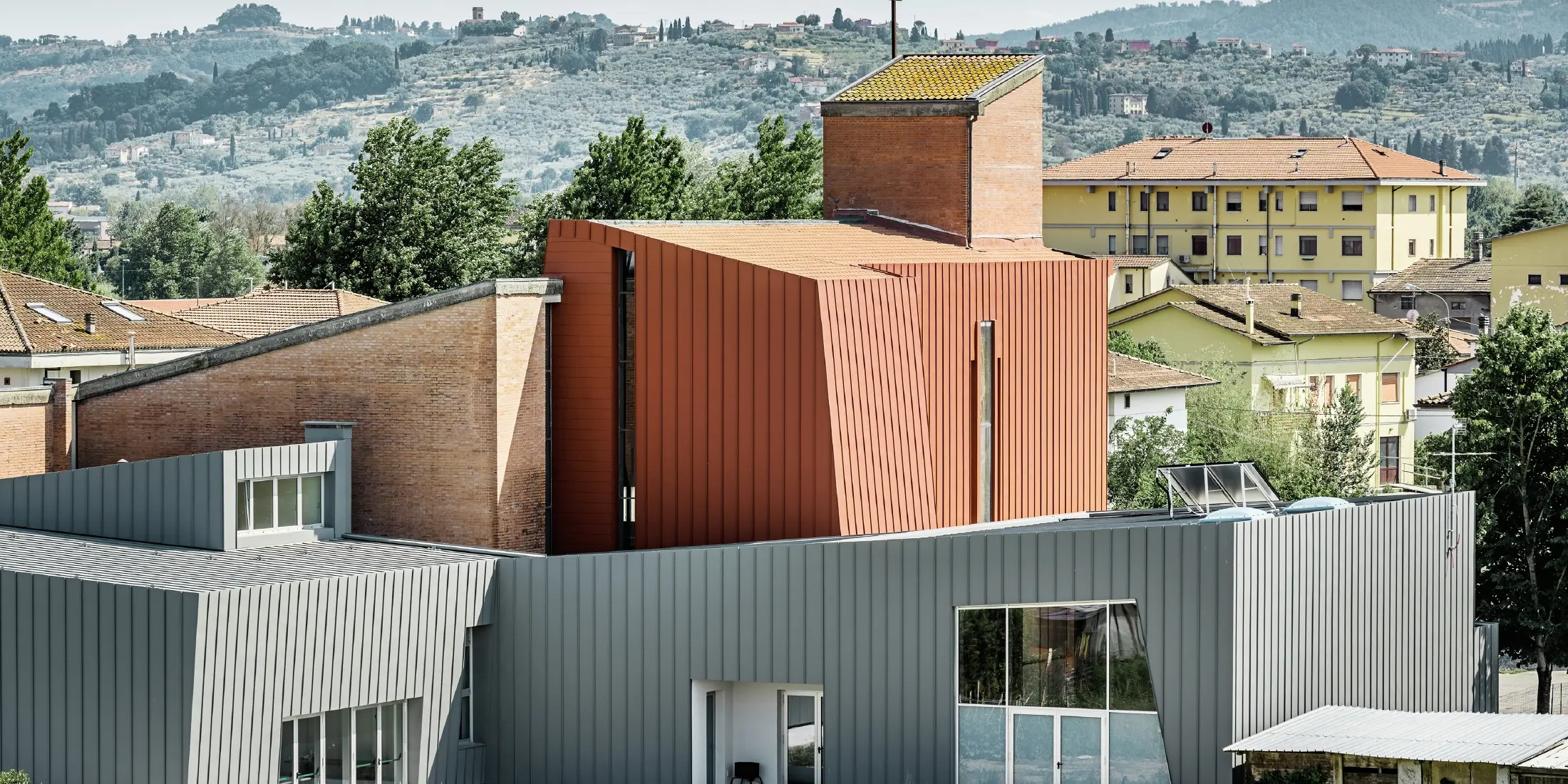 Modernes Kirchengebäude mit markanter Architektur, bestehend aus klaren geometrischen Formen. Der Bau kombiniert hellgraue Aluminiumfassaden mit einem zentralen, ziegelroten Turm aus Metallpaneelen und Ziegelmauerwerk, auf dessen Spitze ein Kreuz steht. Im Hintergrund sind Wohnhäuser und eine hügelige, grüne Landschaft mit Bäumen und Olivenhainen zu sehen.