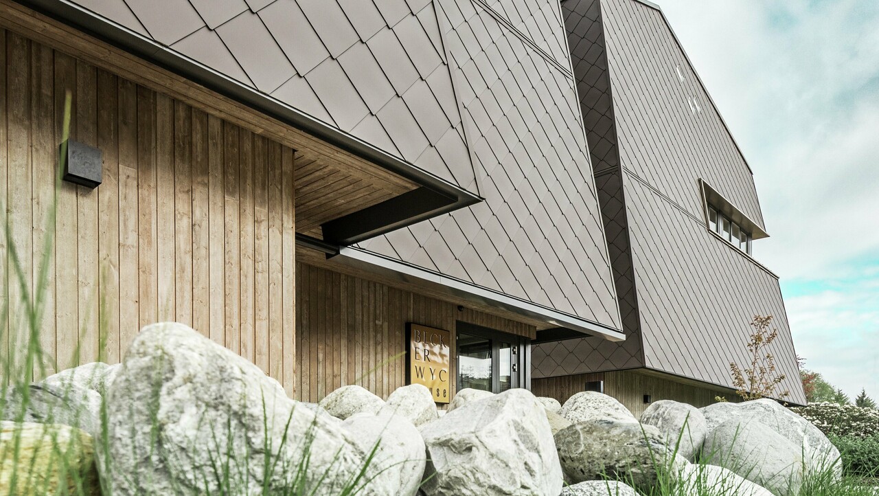 Side view of a hotel, the façade of the ground floor is vertically clad with rather dark wood. Above it is a façade projection, from here the façade is clad with aluminium panels in nut brown.