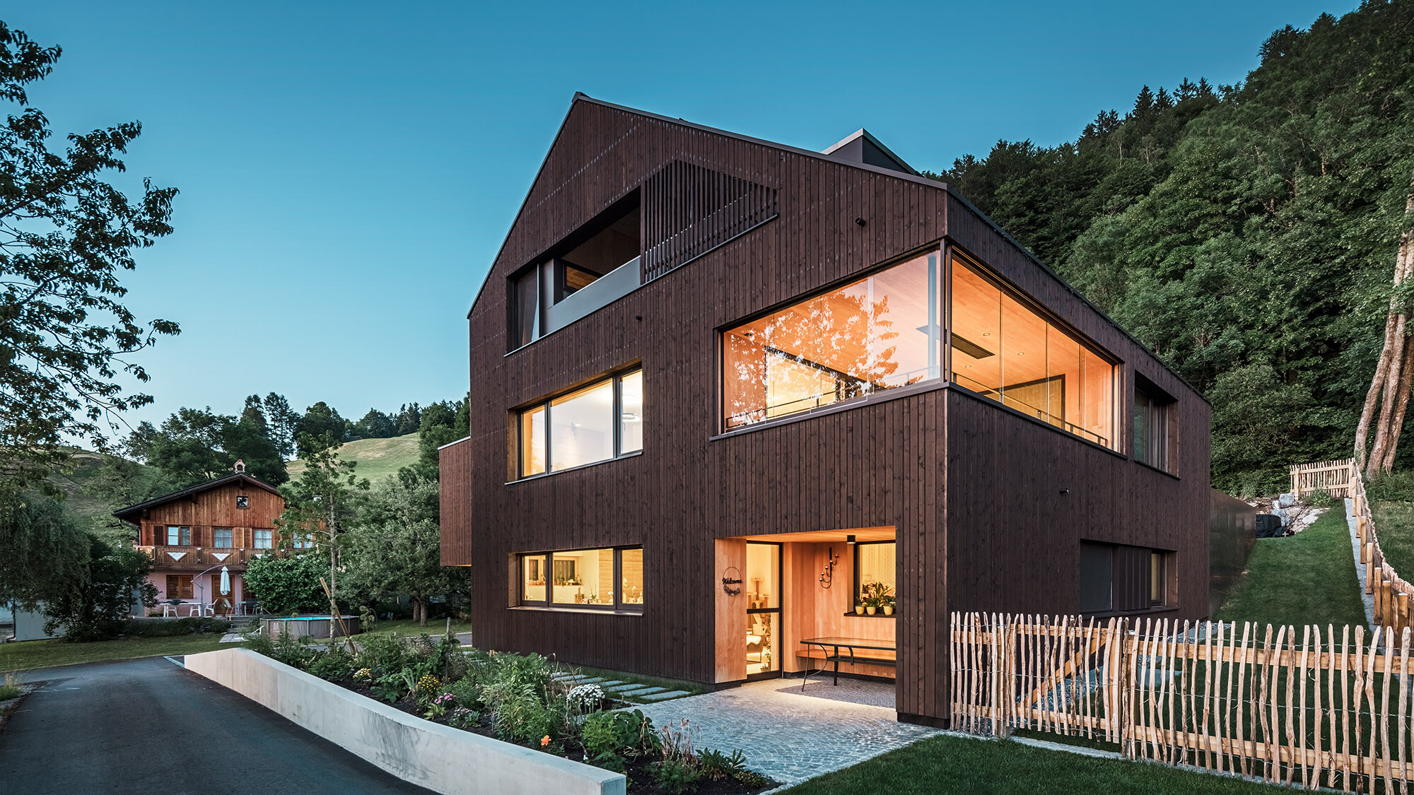 Evening shot of the multi-family home illuminated from the inside with its rural surroundings from a lateral perspective.