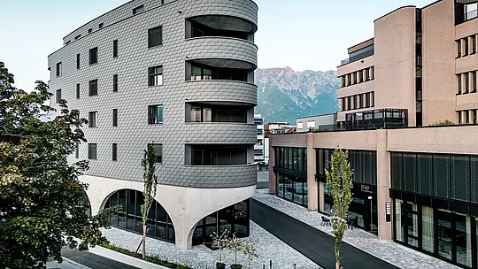 Ansicht des Bernerhauses in Buchs mit markanter, dreieckiger Form und weich gerundeten Balkonen vor alpiner Bergkulisse. Die durchgängige Fassade aus PREFA Wandschindeln in P.10 Hellgrau umhüllt das Gebäude wie eine schuppenartige Aluminiumhaut und erzeugt ein lebendiges Spiel aus Licht und Schatten. Über dem massiven Betonsockel mit eleganten Arkaden wirkt die leichte, witterungsbeständige PREFA Aluminiumfassade besonders präzise, langlebig und wartungsarm – ein architektonischer Eyecatcher im urbanen Umfeld.