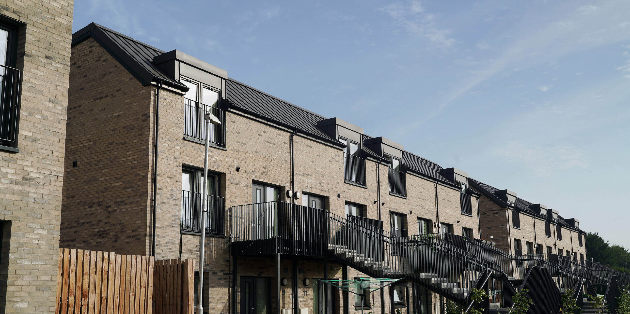 Close-up of a newly built residential complex in Bingham Avenue, Edinburgh, with PREFALZ roofs in P.10 dark grey. The dark aluminium roofs provide an attractive contrast to the light brick walls of the buildings. The individual flats have a balcony or front garden. The staircases are fitted with black banisters, creating a modern and stylish appearance. The front gardens are planted with young trees, which contributes to the aesthetic and ecological quality of the residential complex.