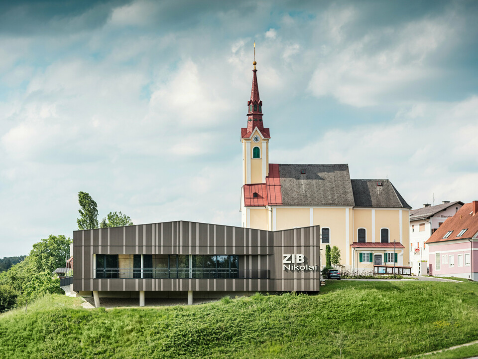 Das Veranstaltungszentrum ZIB Nikolai in St. Veit in der Südsteiermark mit einer modernen, dynamischen Aluminiumfassade in braun-beigegrauen Tönen, im Vordergrund eine grüne Wiese, im Hintergrund die Kirche von St. Nikolai mit gelber Fassade und rotem Dach.