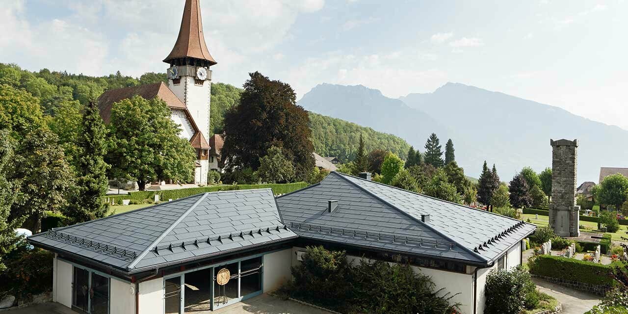 Funeral home with PREFA FX.12 roof panels in anthracite, with a church in the background and the cemetery next to it.