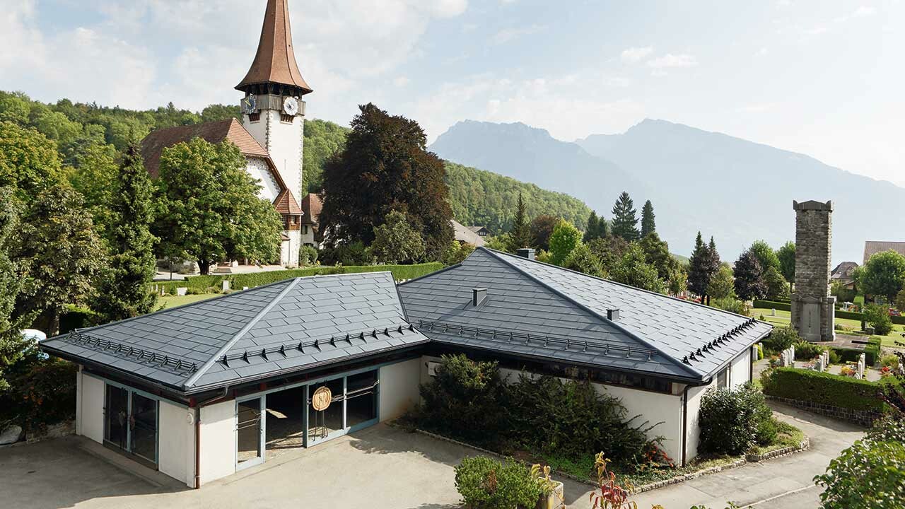 Funeral home with PREFA FX.12 roof panels in anthracite, with a church in the background and the cemetery next to it.