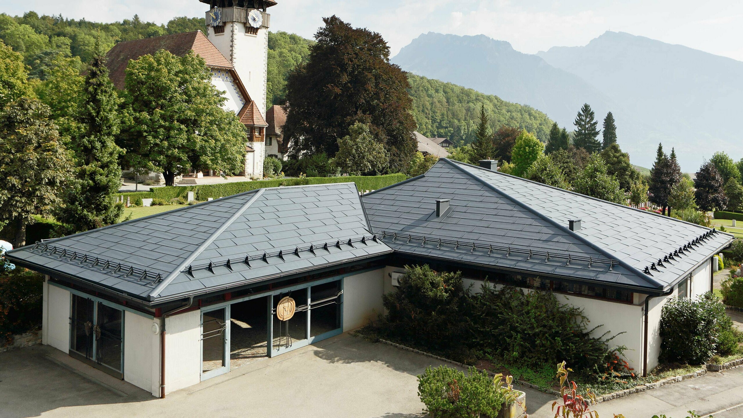 Funeral home with PREFA FX.12 roof panels in anthracite, with a church in the background and the cemetery next to it.