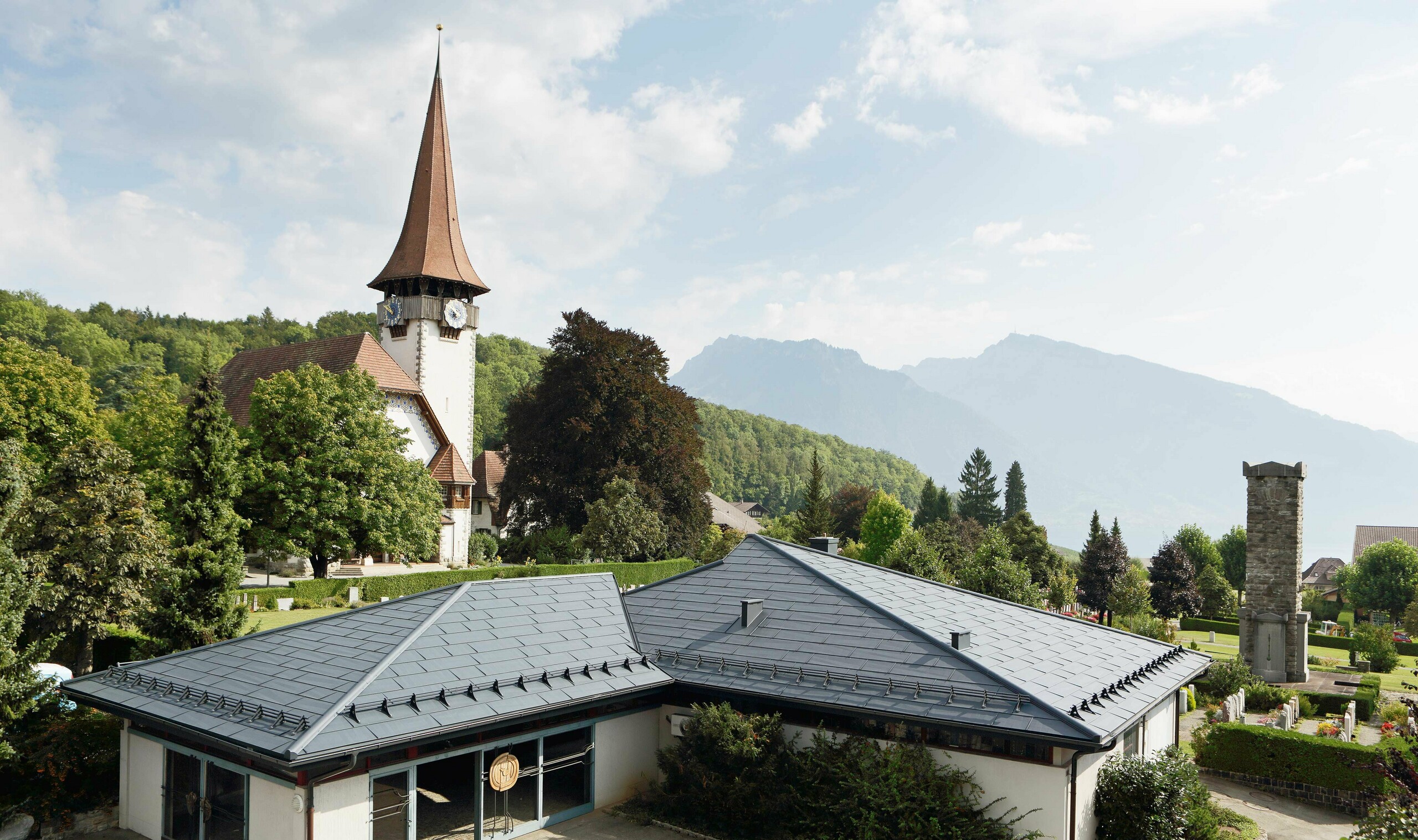 Funeral home with PREFA FX.12 roof panels in anthracite, with a church in the background and the cemetery next to it.