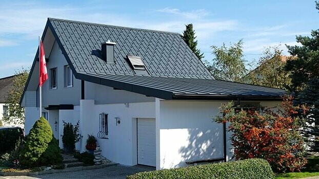 Renovated house with gable roof and adjacent garage. The roof was clad with PREFA roof tiles and the garage with Prefalz in anthracite. Outside the house there is a flagpole with the Swiss flag.