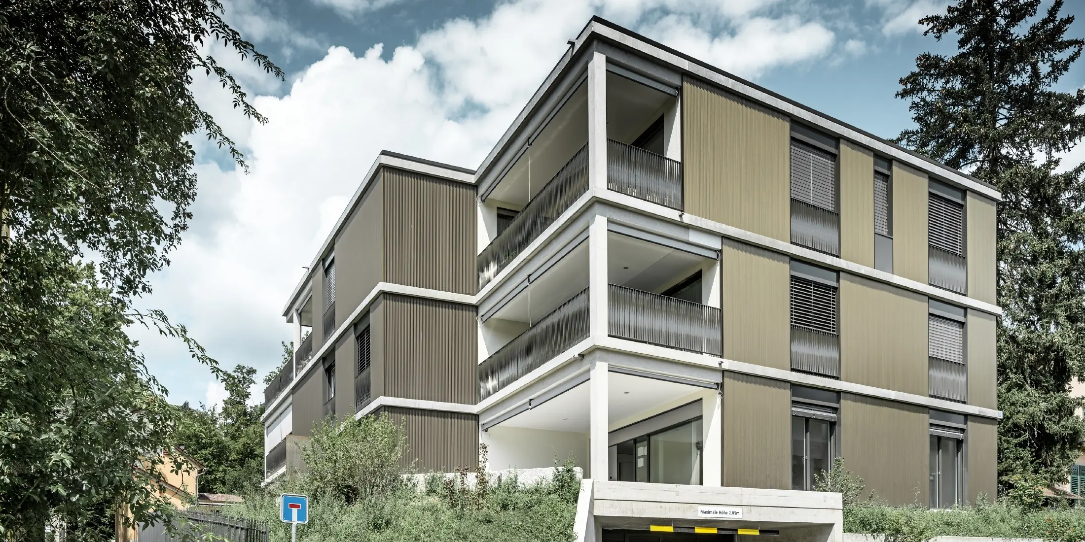 Das Mehrfamilienhaus in Schaffhausen (Schweiz) überzeugt durch seine markante Aluminiumfassade. Die PREFA Fassade aus eloxiertem Zackenprofil in der Farbe Colinal 3165 (E6) macht es zu einem echten Hingucker. Das vertikal ausgerichtete Profil aus stranggepresstem Aluminium unterstreicht die klare Architektur und sorgt für eine optische Kompaktheit des Gebäudes. Die zurückhaltende Farbwahl fügt sich perfekt in das urbane Umfeld ein und verleiht dem Gebäude eine dezente Eleganz. Durchbrochen wird die homogene Gebäudehülle nur von Fenstern, Balkonen, Sichtbeton bei der Garageneinfahrt und einer sichtbaren Säule an der Gebäudeecke.
