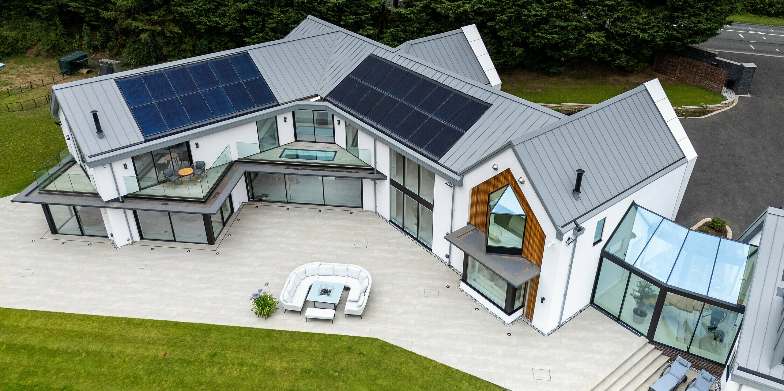 Die Ansicht von oben zeigt die beeindruckende moderne Villa in Draycott In The Clay mit ihrem PREFALZ-Dach in Patinagrau. Das Dach, mit klaren Stehfalzstrukturen und integrierter PV-Anlage, bietet sowohl ästhetische Raffinesse als auch hohe Energieeffizienz. Die große Glasfassade und die markante Holzverkleidung an der Ecke des Hauses heben die architektonische Komplexität hervor. Die weitläufige Terrasse und die gepflegte Gartenlandschaft verleihen dem Gebäude eine harmonische Verbindung zur Natur, während das PREFALZ Aluminiumdach höchsten Schutz und Langlebigkeit garantiert.