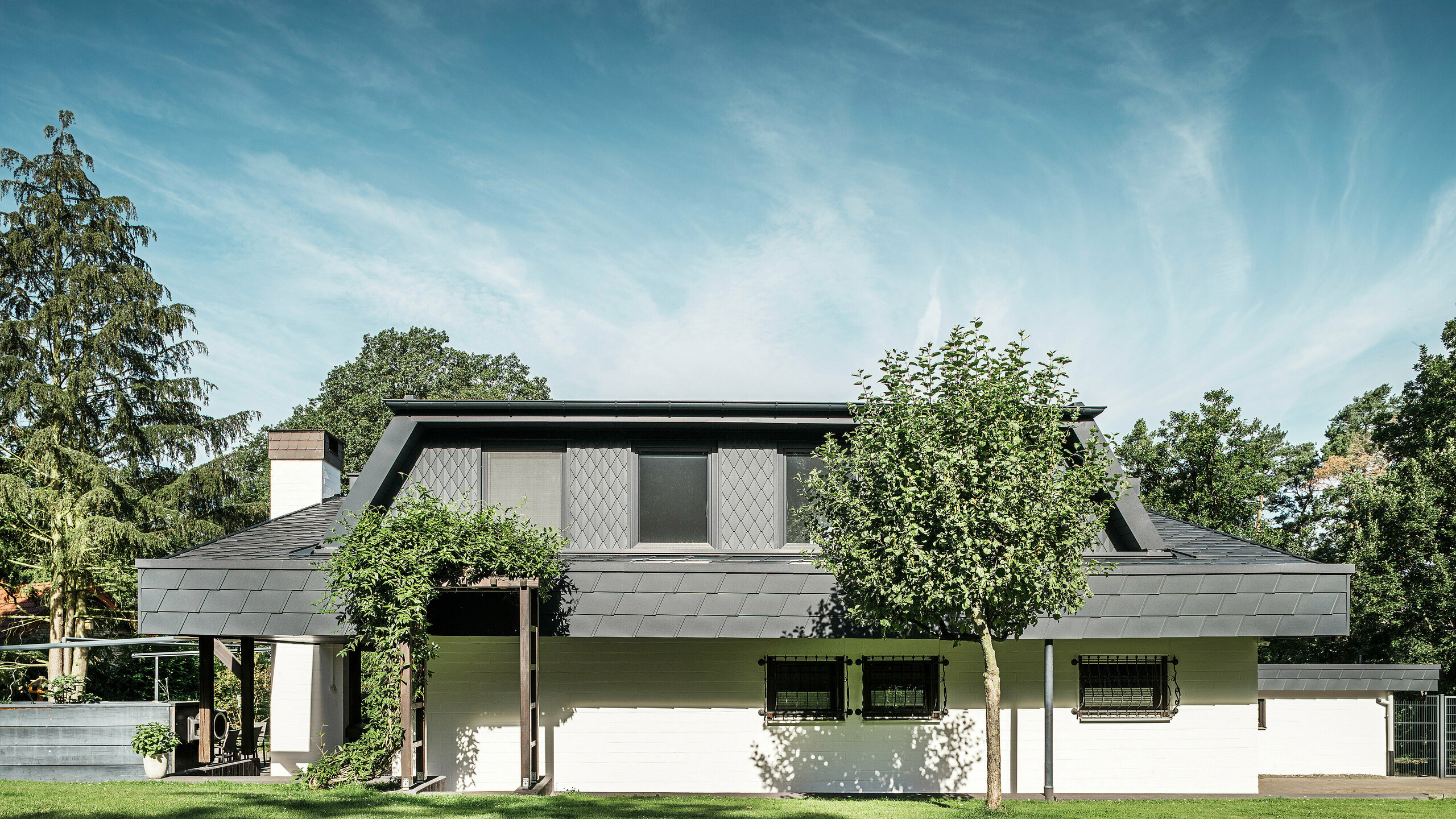 Front view of the garden side of a detached house in Mechernich, featuring a large trapezoidal dormer roof clad with small PREFA P.10 anthracite rhomboid roof tiles. The roof is clad with PREFA DS.19 shingles in the same colour. The façade of the ground floor is painted white, and a green pergola completes the overall look in the garden area. Trees and shrubs frame the view and ensure a harmonious integration into the natural surroundings.