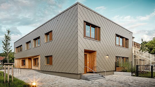 Die moderne Kindertagesstätte in Alzenau beeindruckt mit ihrer klaren Architektur und der hochwertigen PREFA Aluminiumfassade in Bronze. Die Fassade aus Wandrauten 44 × 44 verleiht dem Gebäude eine elegante, strukturierte Optik und verbindet die Funktionsbereiche visuell zu einer harmonischen Einheit. Holzverkleidungen an den Eingängen sorgen für einen warmen, natürlichen Kontrast zum langlebigen Aluminium. Die Kombination aus Massivbau, Holzelementen und vorgehängter hinterlüfteter Fassade steht beispielhaft für nachhaltiges Bauen. Die wartungsarme PREFA Gebäudehülle überzeugt nicht nur durch ihre Recycelbarkeit, sondern auch durch ihre Widerstandsfähigkeit gegenüber Witterungseinflüssen – ideal für zukunftsorientierte Bildungsbauten mit hohem architektonischem Anspruch.
