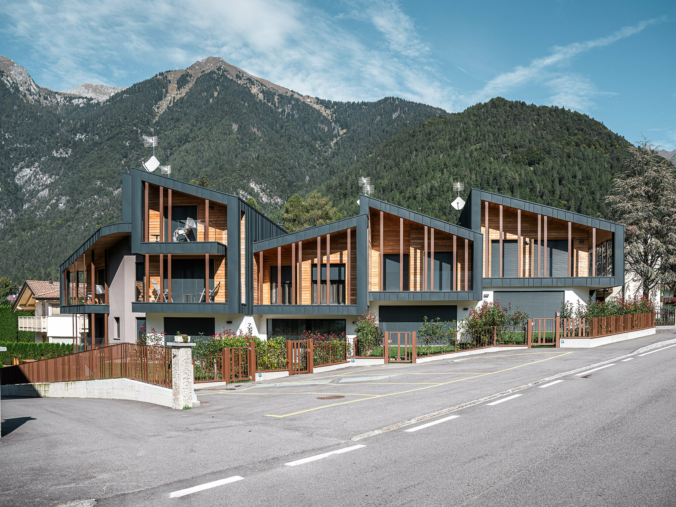 [Translate to English:] Moderne Ferien-Chalets mit anthrazitfarbenen Prefalz-Metalldächern und Holzelementen in einem alpinen Tal, an einer Straße gelegen, mit umzäunten Vorgärten und einem beeindruckenden Bergpanorama im Hintergrund.