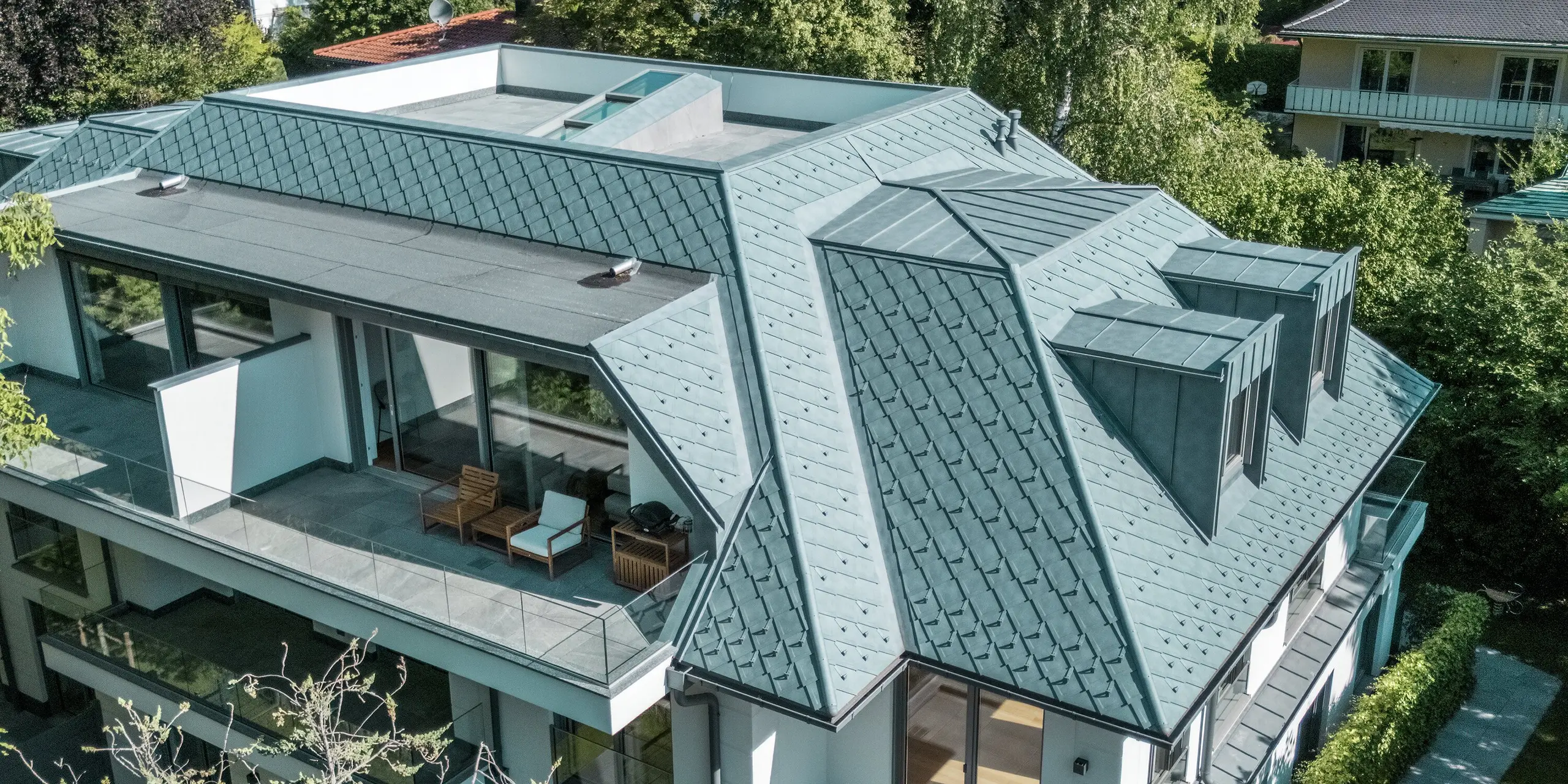 Luftaufnahme eines modernen Mehrfamilienhauses, dessen Dach mit PREFA Dachrauten 29 × 29 in der eleganten Farbe P.10 Steingrau gedeckt ist. Das prägnante, strukturierte Dachdesign wird durch die stilvollen PREFALZ Flachdachgauben ergänzt, die harmonisch in die Architektur integriert sind. Die großzügigen Terrassenflächen und die grüne Umgebung unterstreichen das elegante Gesamtbild. Die Verwendung hochwertiger Aluminiumprodukte sorgt für eine langlebige und witterungsbeständige Lösung, die sowohl ästhetisch als auch funktional überzeugt.
