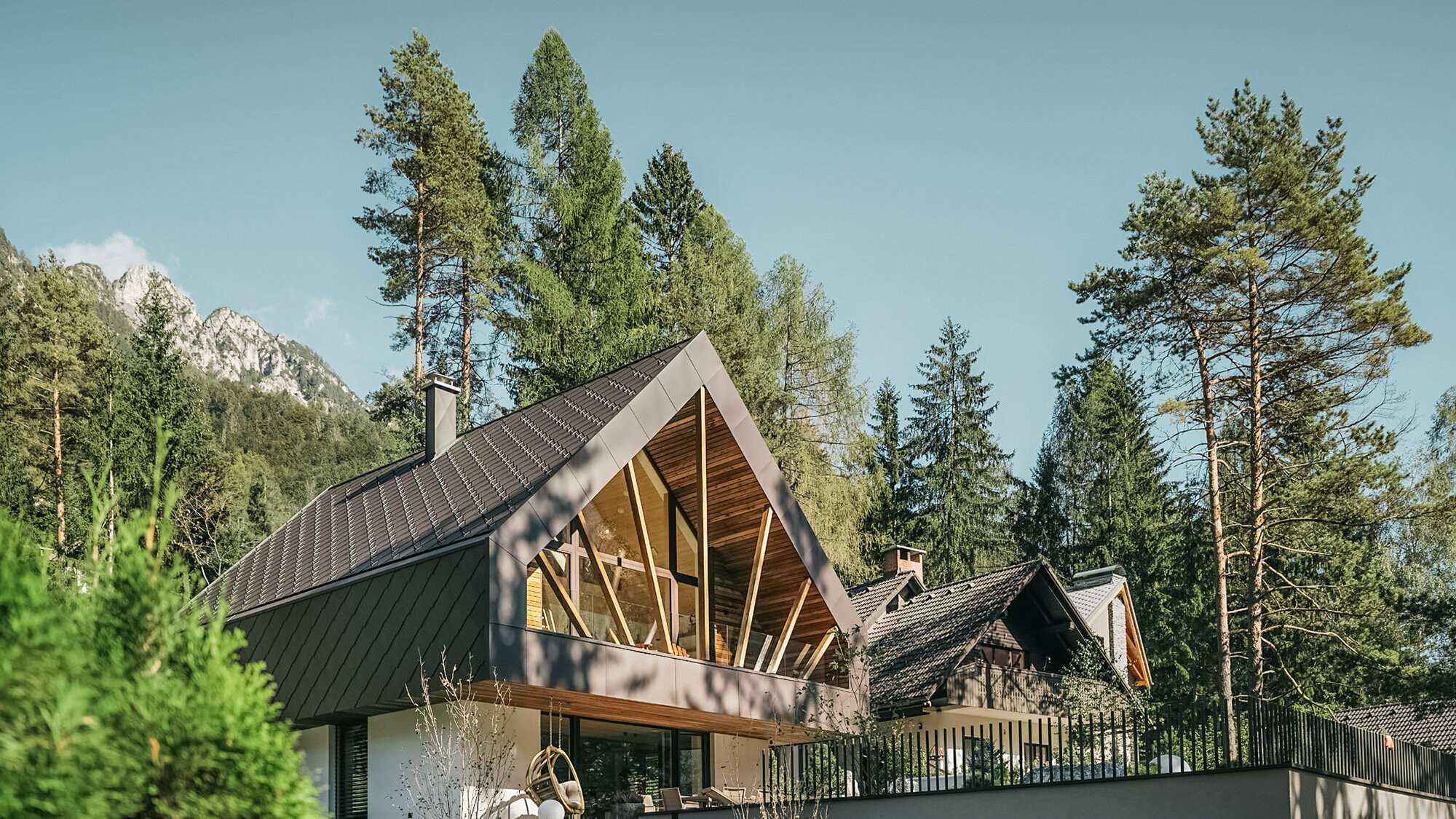 Modern alpine chalet in an idyllic mountain landscape, surrounded by tall pine trees. The house is characterised by a steep, dark pitched roof covered with PREFA roof and wall tiles 44 × 44 in P.10 walnut brown and large, triangular window fronts that allow plenty of light into the interior and provide a view of the surrounding nature. A terrace with railings extends in front of the ground floor.