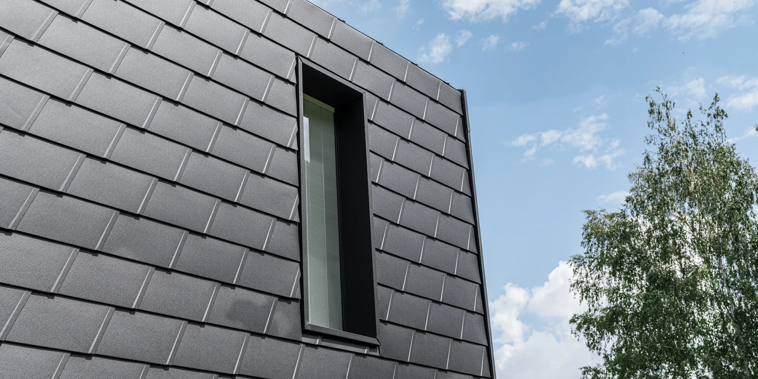 Detailansicht einer Fassade mit PREFA Wandschindeln in Schwarz, die ein schlankes, vertikales Fenster einrahmt. Die Schindeln bieten eine strukturierte, robuste Oberfläche, die ästhetisch mit der natürlichen Umgebung und dem blauen Himmel kontrastiert.