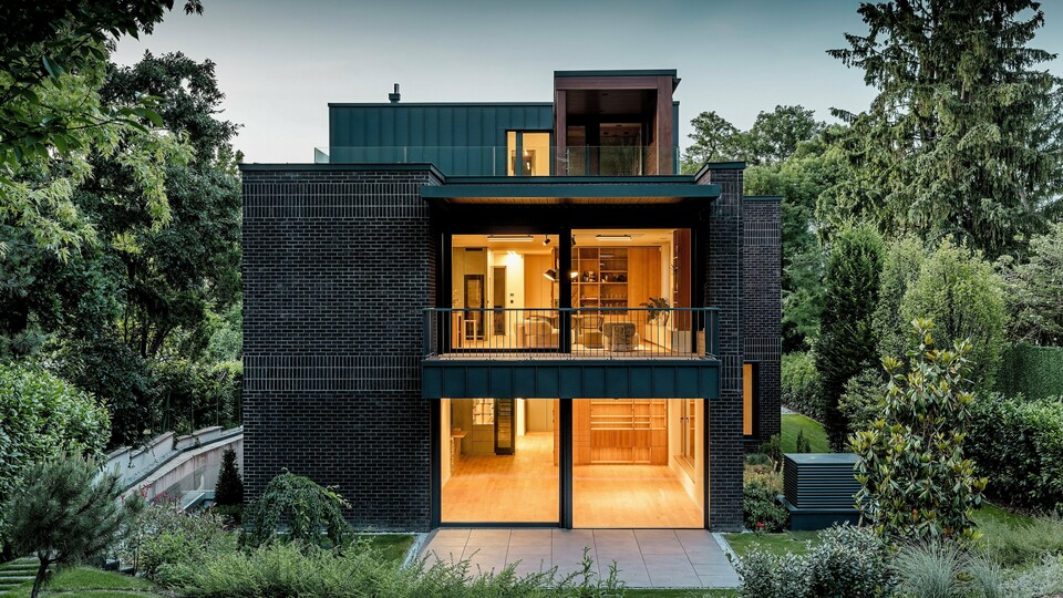 Die Aufnahme zeigt die moderne Rückseite der Budapester Mehrgenerationen‑Villa in der Abenddämmerung. Großzügige Glasfronten öffnen den Blick auf warm erleuchtete Innenräume, während die Kombination aus dunkelroter Backsteinfassade und PREFALZ Aluminium in P.10 Anthrazit klare architektonische Linien betont. Die präzise ausgeführten Winkelstehfalze unterstreichen die hochwertige Verarbeitung und Langlebigkeit der PREFA Aluminiumprodukte. Umgeben von üppigem Grün fügt sich das Gebäude harmonisch in die Gartenlandschaft ein und vermittelt einen eleganten, modernen Charakter.