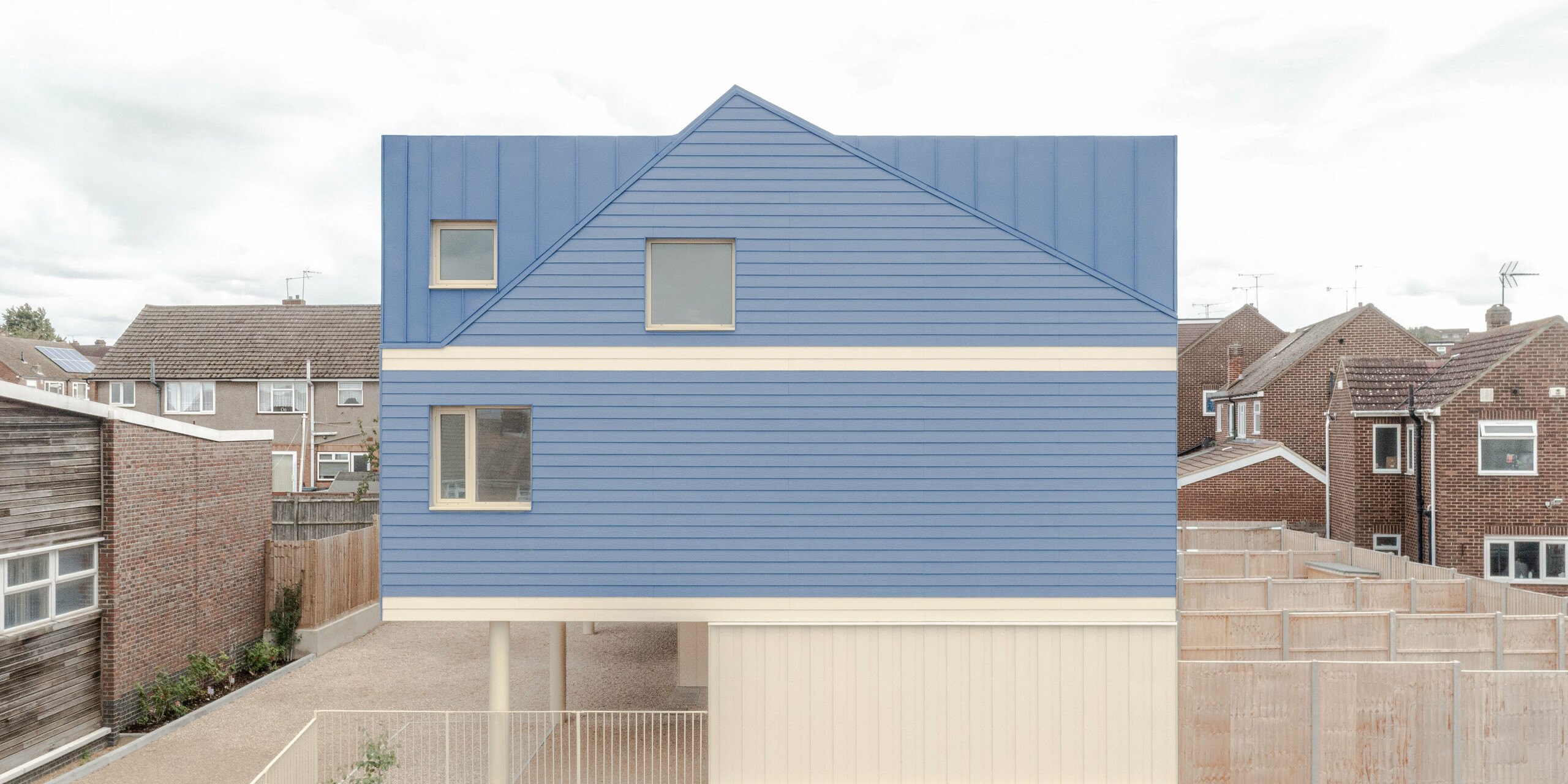 The image shows the striking dusty blue FALZONAL stading seam roof and façade cladding of the Goldsmith Mews terraced housing development, which clearly stands out from the surrounding buildings. The precise folded structures lend depth and rhythm to the aluminium surface, while the light-coloured ground floor areas create a strong, modern contrast. The elevated construction gives a light and open impression. The robust, colour-stable PREFA aluminium offers long-lasting weather protection, high dimensional stability and enables a clear, bold architecture that gives the building ensemble an unmistakable character.