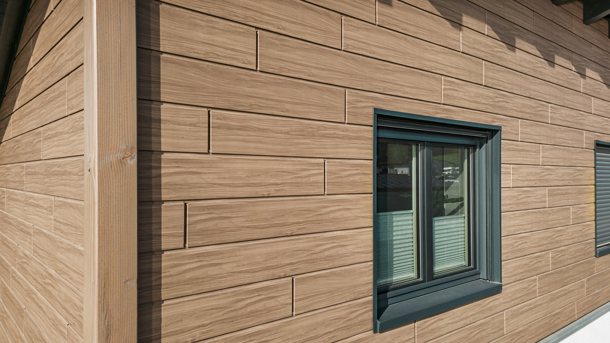 Detailaufnahme der Gebäudekante am Obergeschoss eines Einfamilienhauses in Bruck. Das obere Stockwerk ist mit einer Fassade aus Aluminium in einer Holzoptik verkleidet. Die PREFA Sidings in Walnuss Braun bilden einen harmonischen Übergang zum weißen Gebäudesockel.