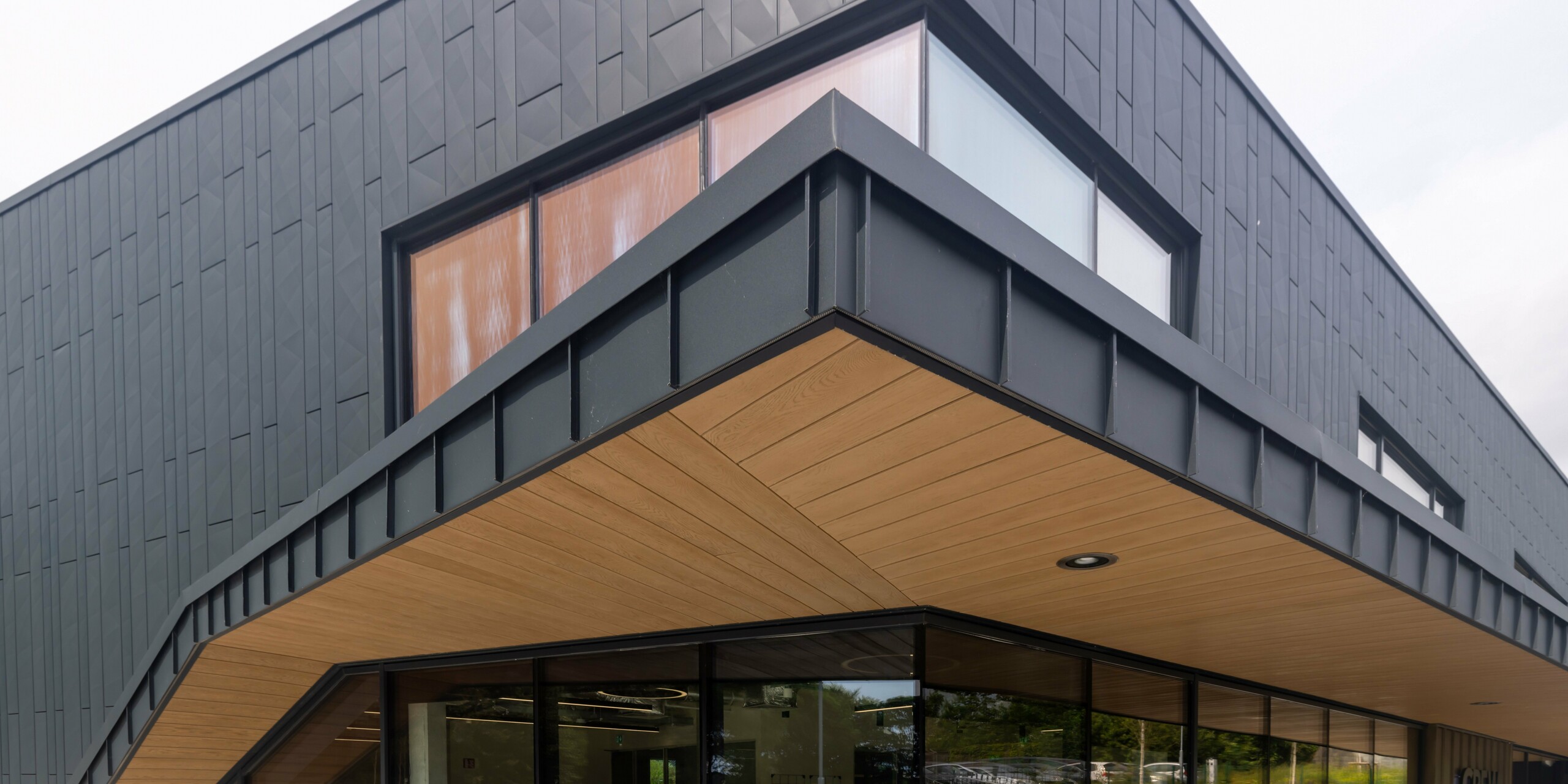 Detailed view of the entrance area of the CREW building in Galway, Ireland, with modern architecture and high-quality PREFA sheet metal façade. The façade consists of Siding.X façade panels and PREFALZ standing seam cladding in the colour P.10 anthracite. The canopy with an elegant wood look on the underside complements the anthracite-coloured aluminium cladding and gives the building a warm, inviting atmosphere. The large window areas ensure bright and open interior spaces.