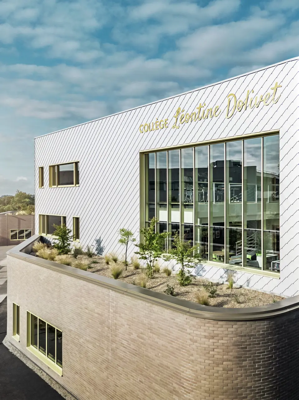 Das Foto zeigt eine markante Ecke des Collège Léontine Dolivet in Cesson-Sévigné. Die Fassade besteht aus silbermetallischen PREFA-Wandelementen, die diagonal angeordnet sind und das Licht reflektieren. Besonders auffällig ist die geschwungene Form des Obergeschosses, die dem Gebäude eine sanfte und moderne Anmutung verleiht. Große Glasflächen geben den Blick ins Innere frei, während gelbe Fensterrahmen farbliche Akzente setzen. Im Vordergrund befindet sich eine bepflanzte Fläche mit Kies und Sträuchern, die die Architektur harmonisch ergänzt. Der leicht bewölkte Himmel hebt die Metalloberfläche zusätzlich hervor.