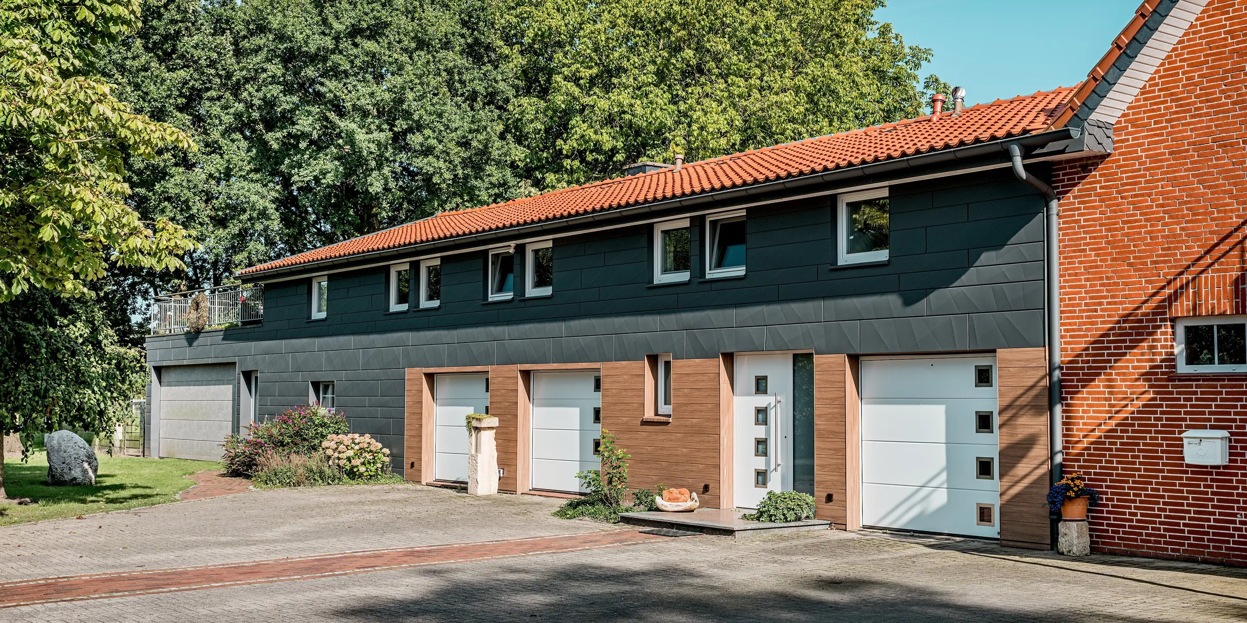 Modern saniertes Einfamilienhaus mit markanter PREFA Aluminiumfassade in horizontaler Gliederung. Eyecatcher sind die Kombination aus PREFA Siding in warmer Walnussbraun-Holzoptik im Sockel- und Eingangsbereich sowie Siding.X in P.10 Dunkelgrau im Obergeschoss. Die präzisen Kantungen der dunklen Paneele erzeugen spannende Licht- und Schattenspiele und verleihen der Fassade eine zeitgemäße Eleganz. Weiße Garagentore und klar gefasste Fenster setzen bewusste Kontraste. Die langlebigen, witterungsbeständigen und pflegeleichten PREFA Aluminiumpaneele eignen sich ideal für nachhaltige Sanierungen und verbinden modernes Design mit hoher Funktionalität.