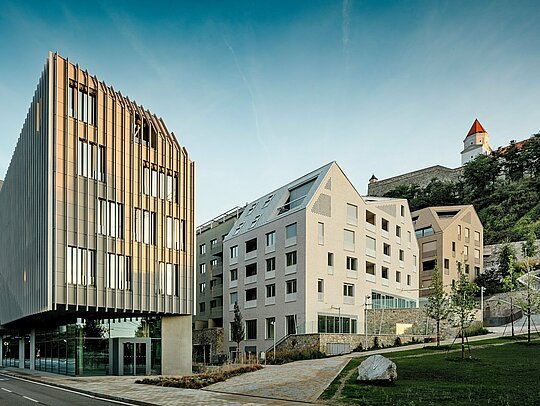 Markantes Zusammenspiel aus moderner Architektur und historischem Kontext im Stadtquartier Vydrica in Bratislava: Im Vordergrund fasziniert ein skulpturaler Baukörper mit vertikaler Fassadenstruktur, während dahinter steil geneigte Dächer mit PREFA Aluminium Dachschindeln in Prefaweiß und Sandbraun präzise Akzente setzen. Die klar gegliederten Dachflächen mit integrierten Fenstern bilden einen starken visuellen Kontrast zur Burgkulisse am Hang. PREFA Aluminium überzeugt durch hohe Formbarkeit, Langlebigkeit und extreme Witterungsbeständigkeit – ideal für architektonisch anspruchsvolle Stadtprojekte.