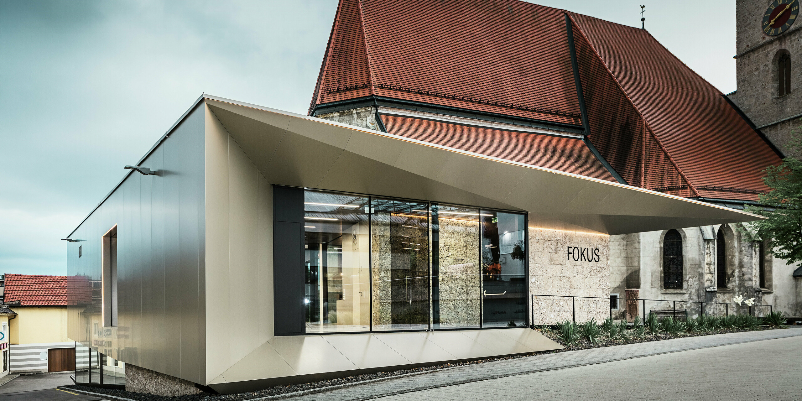 Im Vordergrund des Pfarrheims in Sierning glänzt die Aluminiumfassade durch ihre saubere Linienführung und hohe Formstabilität. Die Kombination aus Glasflächen und bronzefarbenen PREFA Aluminium Verbundplatten betont Offenheit, Beständigkeit und gestalterische Präzision bei gleichzeitiger Integration ins historische Umfeld.