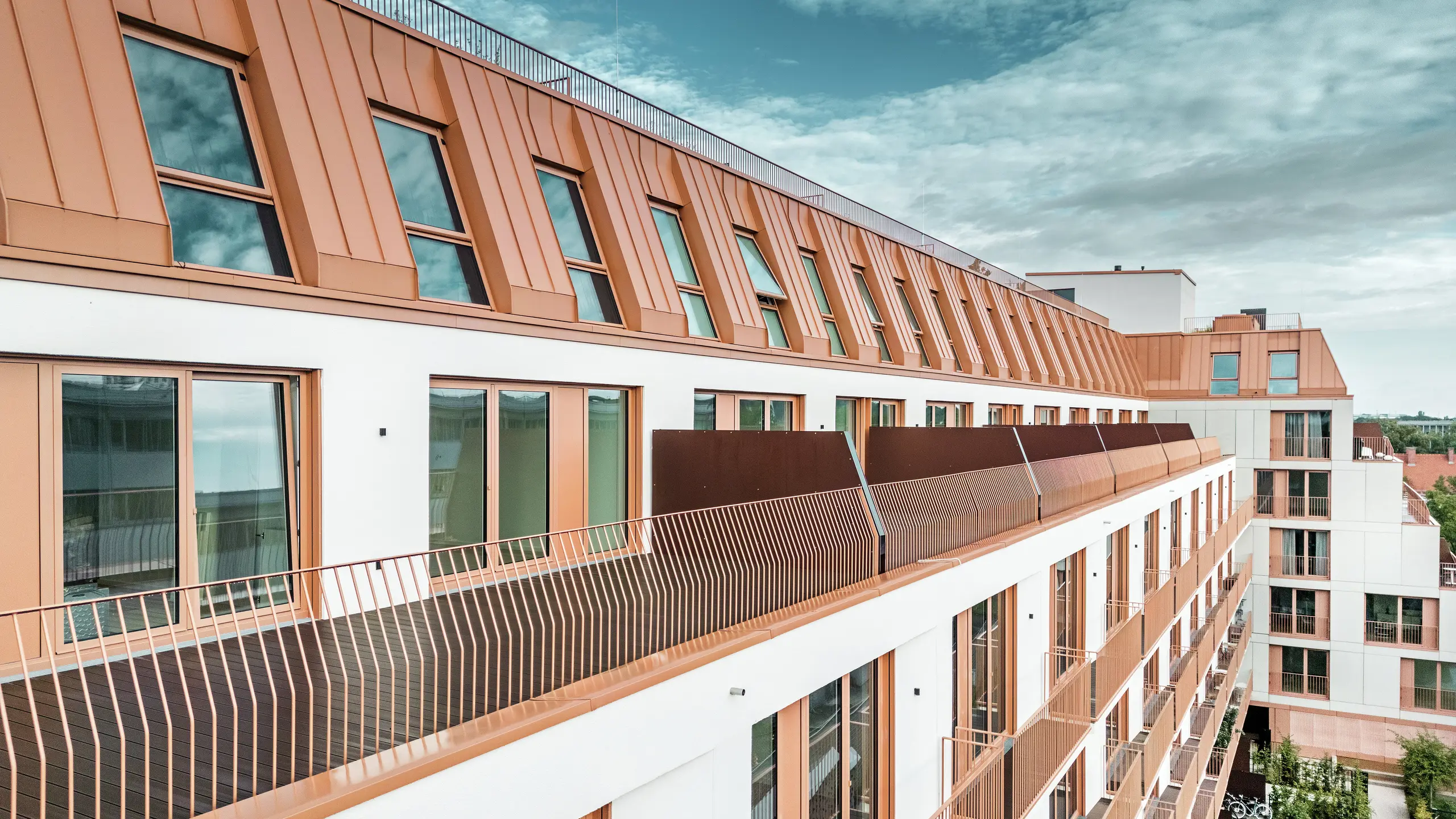 Modern residential building Van B in Munich with striking roof landscape made of PREFALZ aluminium in the special colour IGP Orange HDP. The durable and low-maintenance roof covering from PREFA ensures aesthetic unity and high weather resistance. The elegant façade design with copper-orange window frames and generous balcony areas underlines the innovative architectural concept. The ideal solution for urban construction projects with high standards of design and functionality.