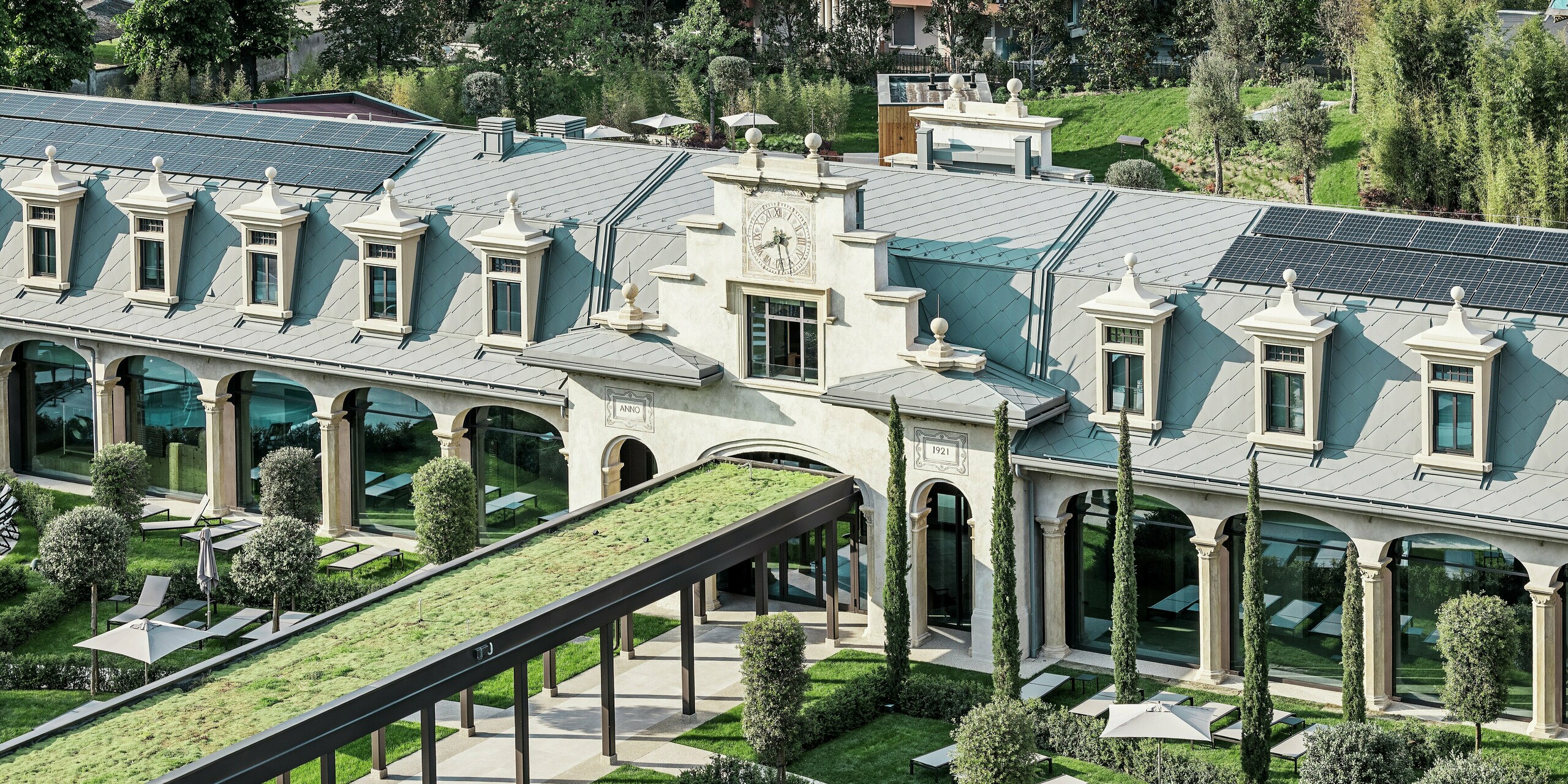 Dieser elegant restaurierte Jugendstilbau im Mailänder Stadtteil San Siro besticht durch seine markante Dacheindeckung aus PREFA Dachrauten 44 × 44 sowie PREFALZ Stehfalzbahnen unter der großflächigen Photovoltaikanlage. Beide Aluminiumprodukte sind in der Farbe P.10 Hellgrau gehalten. Im Vordergrund befindet sich ein begrünter Laubengang, der von einer gepflegten Gartenanlage mit Liegeflächen, schattenspendenden Sonnenschirmen, Zypressen und geschwungenen Wegen umgeben ist, die für eine maximale Aufenthaltsqualität sorgen.