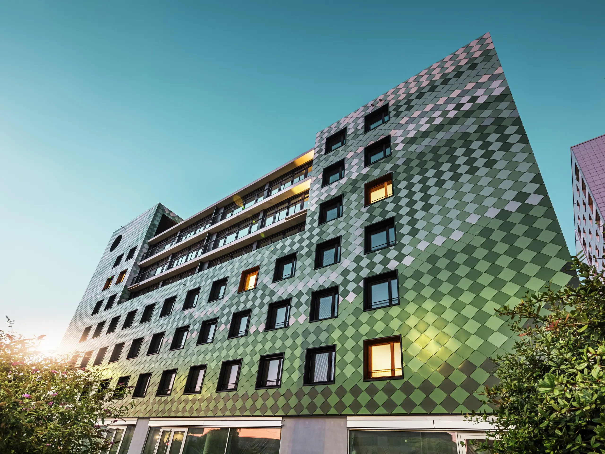 The modern residential and commercial building ‘Pick-Nick’ in Dresden has a striking PREFA façade made of aluminium rhomboids in the special colours olive green, grey green, reseda green, opal green, light ivory, pearl white and light bronze. The creative design of the façade is reminiscent of a rice field and creates a lively play of colours. The clear architecture, the black window frames and the durability of the PREFA materials characterise the building. The low sun is reflected on the façade and the window surfaces.