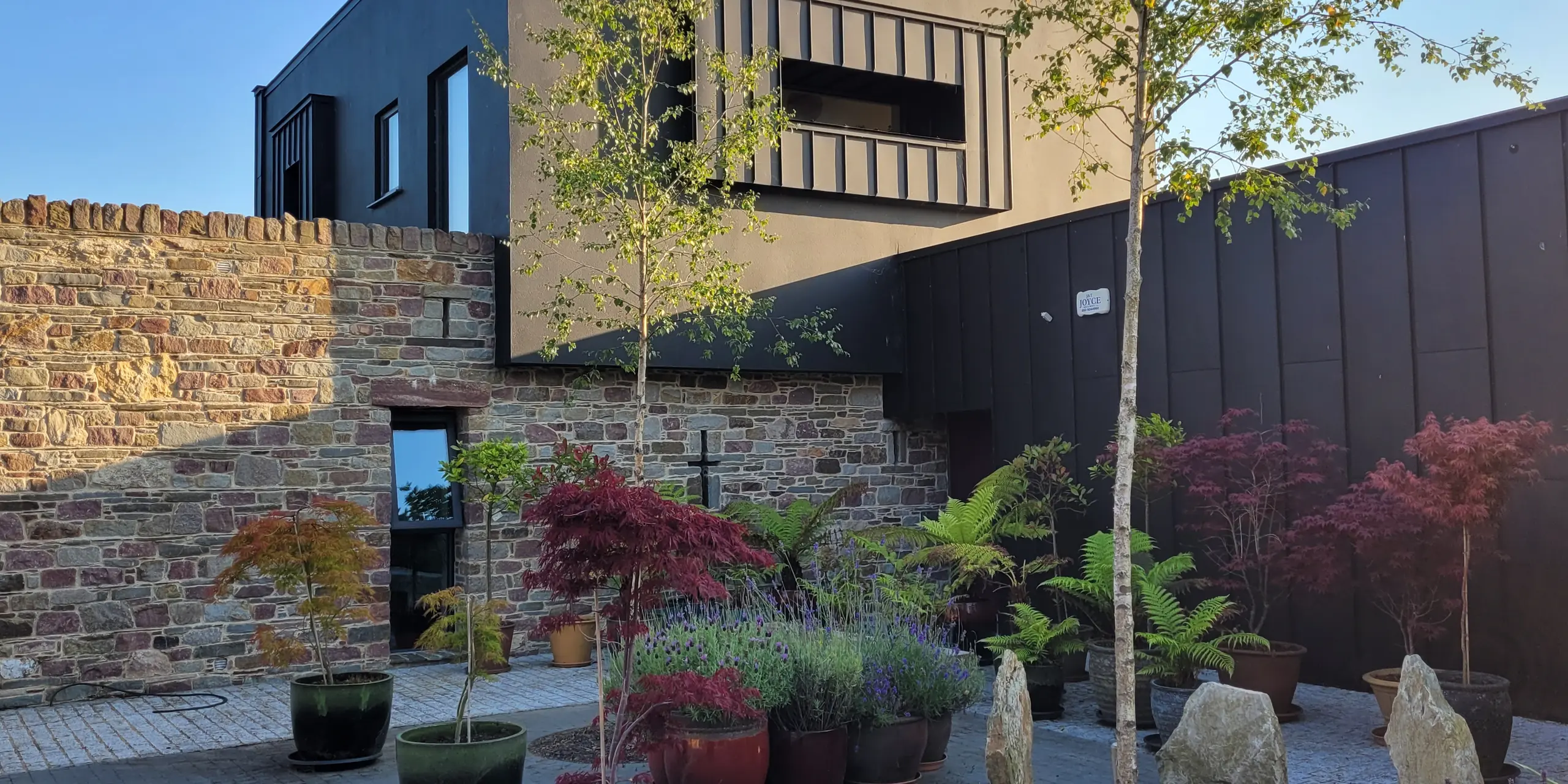 Das Bild zeigt das moderne Architektenhaus in Wexford. Dessen Fassade aus PREFALZ in der Farbe P.10 Schwarz bildet einen starken Kontrast zur warmen Natursteinwand. Die klaren Aluminiumflächen reflektieren das goldene Abendlicht und betonen die präzise Linienführung der Konstruktion. Im Vordergrund schaffen sorgfältig platzierte Pflanzen und Ziersteine eine ruhige, beinahe meditative Atmosphäre. Der japanische Ahorn unterstreicht die hochwertige Pflanzenauswahl. Die robuste und witterungsbeständige PREFA-Fassade verleiht dem Gebäude nicht nur Charakter, sondern auch langlebigen Schutz und architektonische Tiefe.