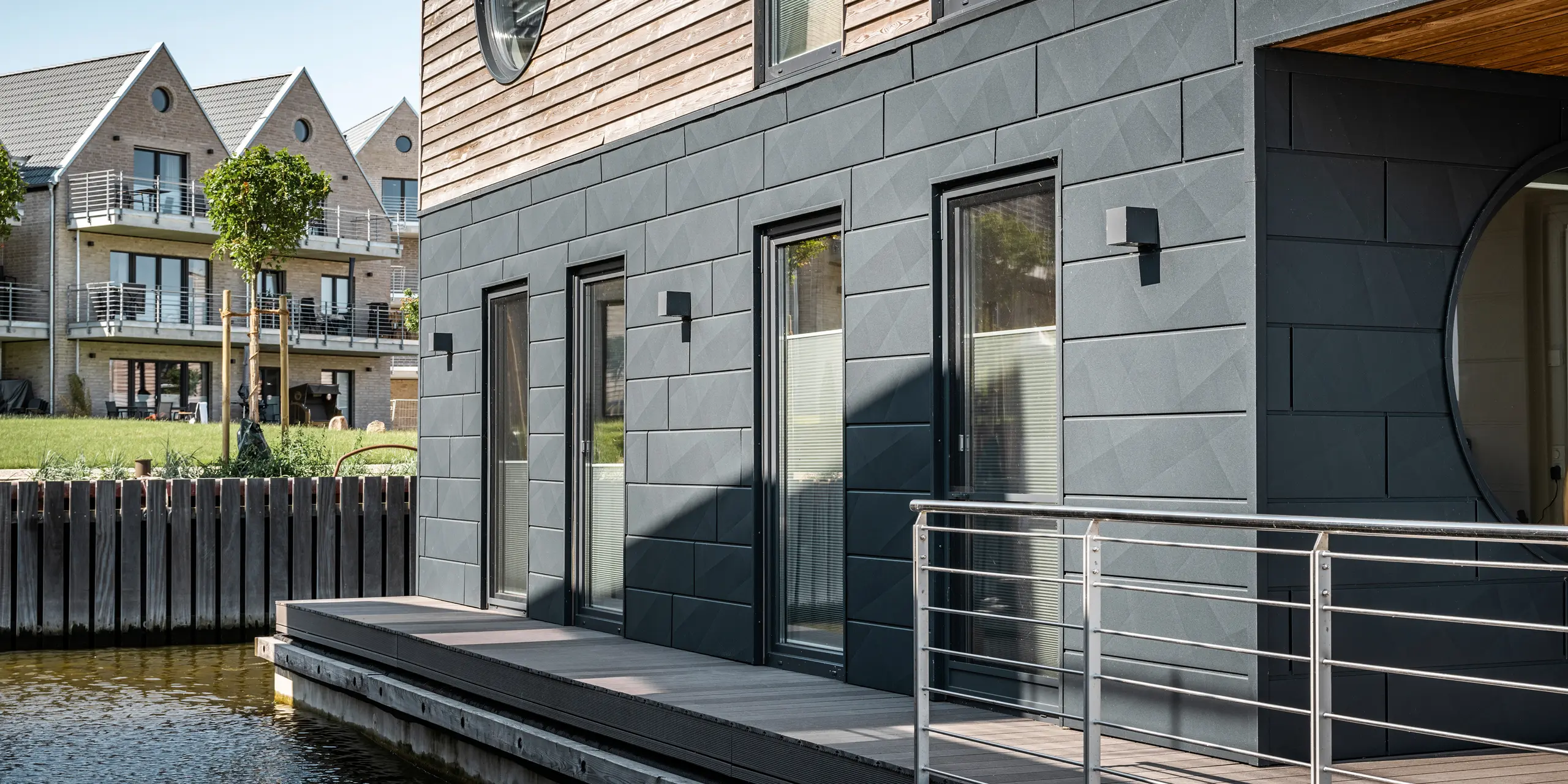 Detailaufnahme eines Wasserhauses im Pionierhafen Schleswig, das die hochwertige PREFA Siding.X Fassade in P.10 Anthrazit zeigt. Das Zusammenspiel von moderner Aluminiumverkleidung und natürlichen Holzelementen spiegelt eine Symbiose aus Funktionalität und Design wider. Im Hintergrund ergänzen traditionelle Häuser mit Giebeldächern und begrünten Außenbereichen die Szenerie und betonen das harmonische Zusammenspiel von alter und neuer Architektur am Wasser.